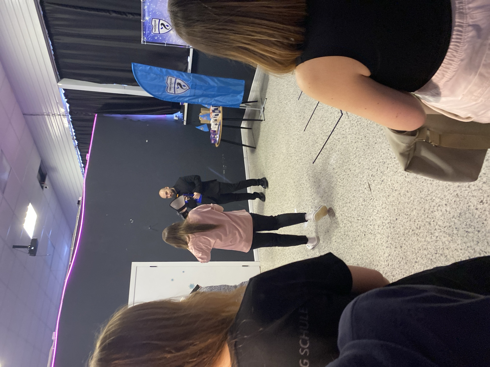 The scene is an indoor event possibly in Chaikovsky, Russia, given the presence of a blue flag with a logo and text in Russian. A stage is visible with a black backdrop, and what seems like a display table. A man in a black suit is holding a blue object, perhaps performing. A woman in a pink shirt and black pants stands near him. People, partially visible, are in the foreground, likely the audience. The room is lit with overhead lighting, some of which have a pinkish hue. The floor is speckled.