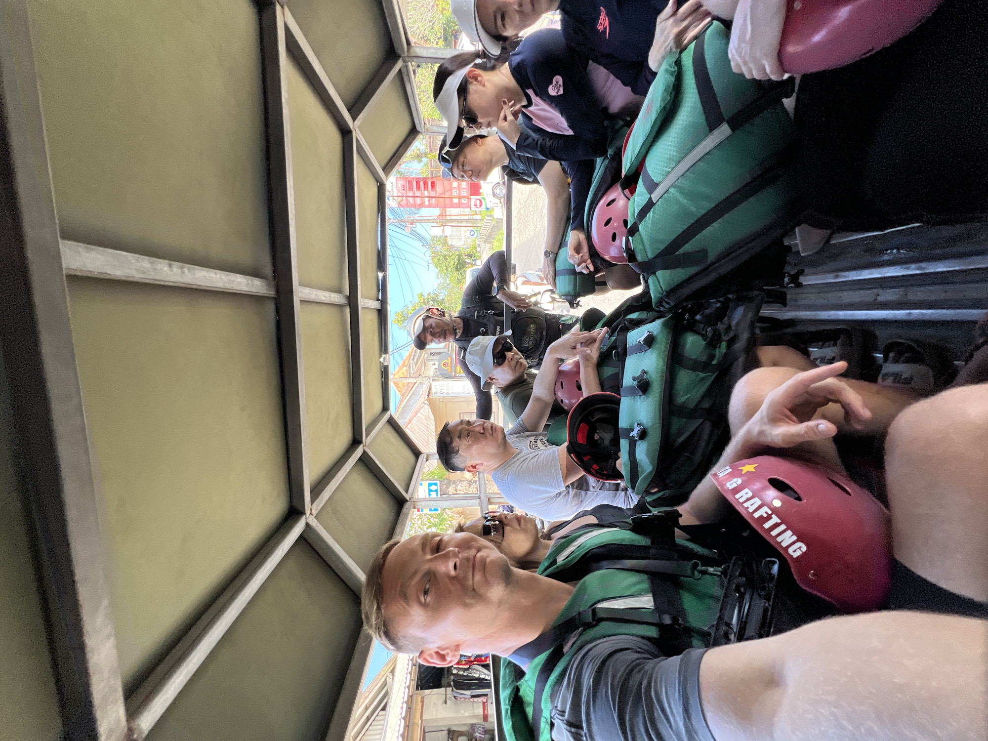 Captured from the vantage point of a moving vehicle, possibly a truck or bus, the image presents a group of people in Ubud District, Indonesia. The passengers appear to be tourists or participants in an outdoor activity, likely white water rafting, as indicated by the presence of life vests and helmets bearing the word "RAFTING." The photograph seems to have been taken by one of the individuals within the group using a camera phone. Other passengers visible include women wearing hats, and some wearing sunglasses. The surrounding environment reveals the scenery of Ubud, characterized by buildings, roads, and vegetation visible through the vehicle's opening. The lighting indicates a bright, sunny day.