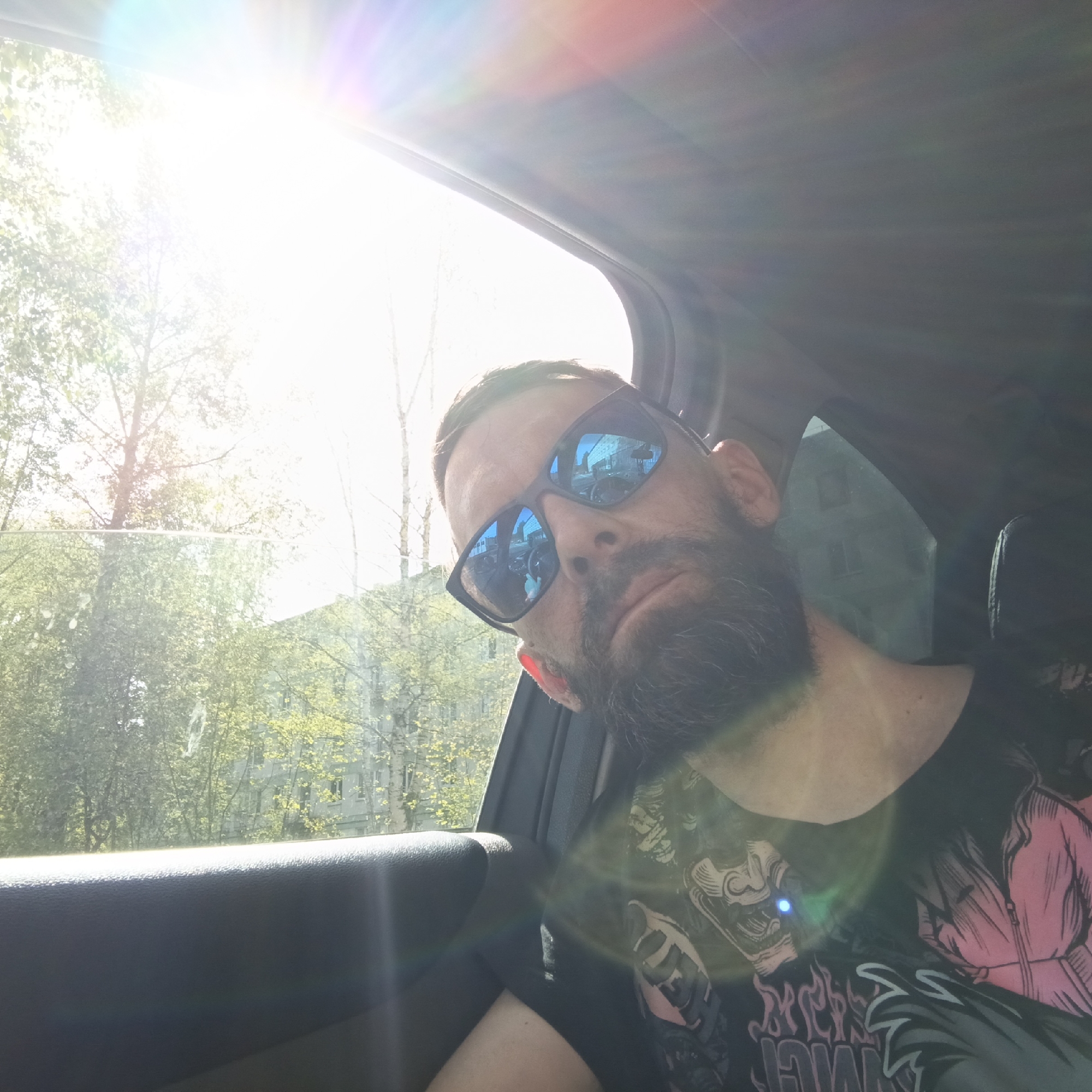 A man with a beard and sunglasses is sitting in a car. The background is a blurred view of trees and a building. The photo was most likely taken during the day. The photo was taken in the city of Chaikovsky, Russia.
