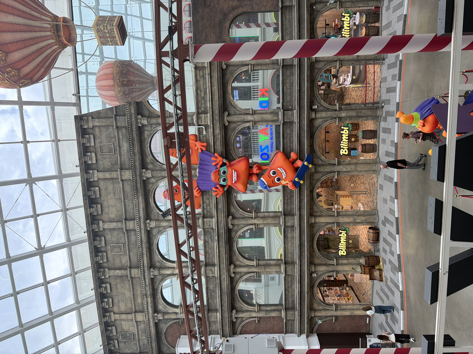 The image shows the interior of a large, multi-level shopping mall with a glass ceiling. The architecture of the mall is designed to resemble old European buildings with stone facades, arched windows, and columns. Decorative elements include oversized cartoon characters hanging from a walkway, hot air balloon decorations suspended from the ceiling, and large candy-themed displays in shop windows. People are seen walking on the ground level and on a balcony. The shops visible have signage that says "Bilimbi". The overall impression is of a brightly lit, themed entertainment and shopping environment.