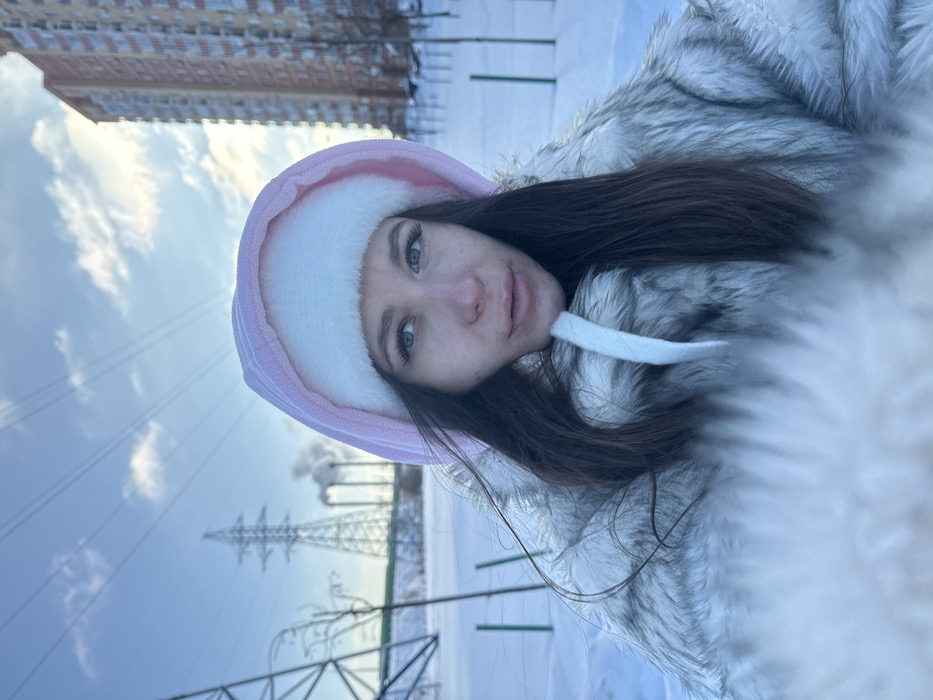 The image captures a female individual outdoors in a snowy environment in Molodi, Russia, under a clear blue sky with scattered clouds. The woman is positioned centrally, viewed in a close-up, selfie-style shot. She has long, dark brown hair and is wearing a light gray or white faux fur coat. Over the coat, she wears a light pink hooded garment featuring a white fluffy lining around the hood's opening, with a white drawstring visible near her chest. Her facial expression is neutral, with her gaze directed slightly towards the right of the frame. The background on the left reveals two large metal electricity pylons and multiple power lines, beyond which industrial chimneys are visible emitting white smoke. The ground in the foreground and mid-ground is covered uniformly in snow, with sparse bare vegetation and several small green posts or markers protruding from the snow. On the right side of the background, a tall, multi-story residential building with orange and beige paneling stands amidst the snow and bare trees.