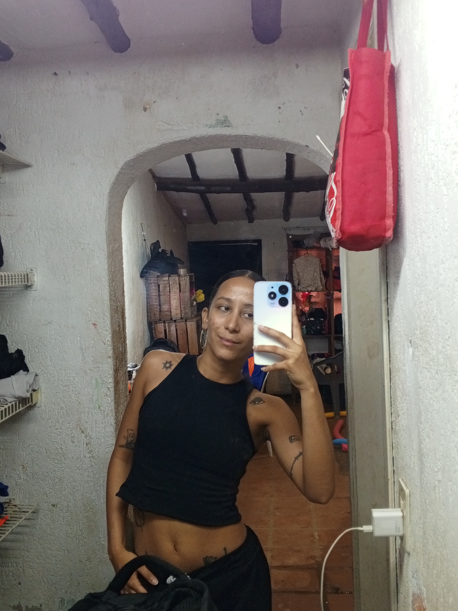 A medium shot features a light-skinned person taking a selfie in a mirror. The person is wearing a black tank top and black pants. Tattoos are visible on their arms, shoulder, and abdomen. They are holding a white smartphone with three camera lenses. The mirror reflects a hallway or entryway with an arched opening and a wooden-beamed ceiling. Behind the person in the mirror is a room with shelves, clothing, and various items. A red tote bag hangs on the wall to the right of the mirror. The walls are textured and painted white, with signs of wear and tear. An electrical outlet and phone charger are visible on the wall next to the mirror. The photograph was taken indoors in Porlamar, Venezuela.