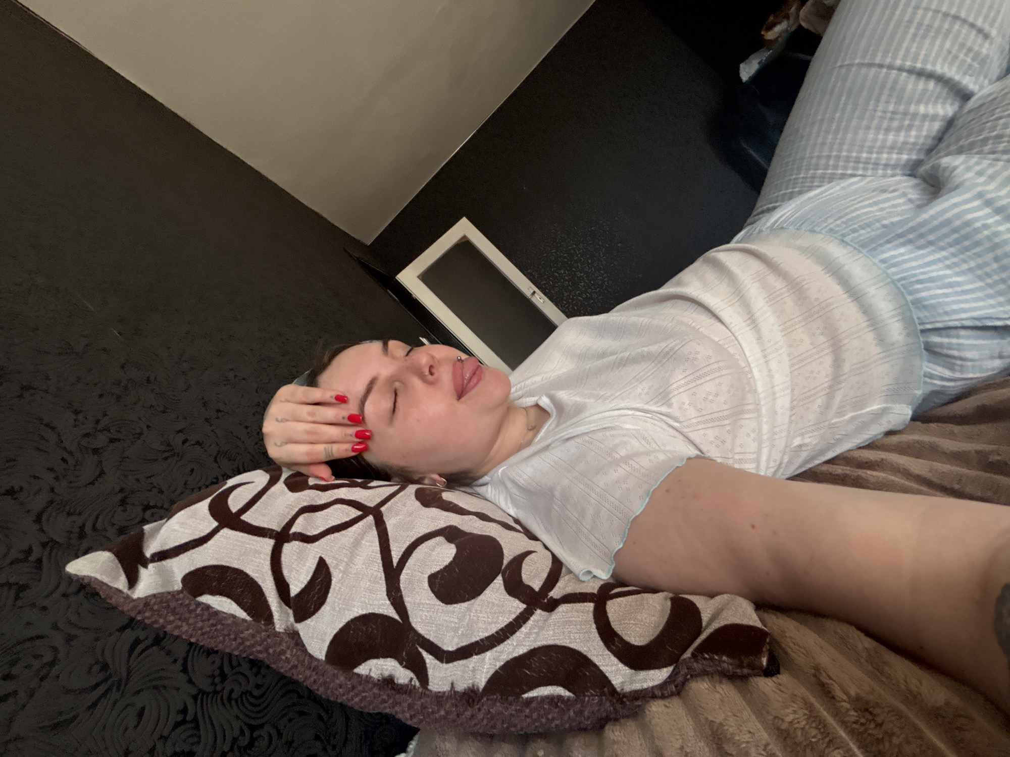 A woman is lying on a bed with a patterned pillow under her head. She is wearing a white, patterned shirt and light blue striped pajama pants. Her left hand rests on her forehead, and her nails are painted red. She has multiple piercings and her eyes are closed, her tongue sticking out slightly. The bed has a brown, textured blanket. The background includes a black wall with a subtle pattern, a white door or frame, and part of a white wall or ceiling. The location and the time are not discernible from the image.