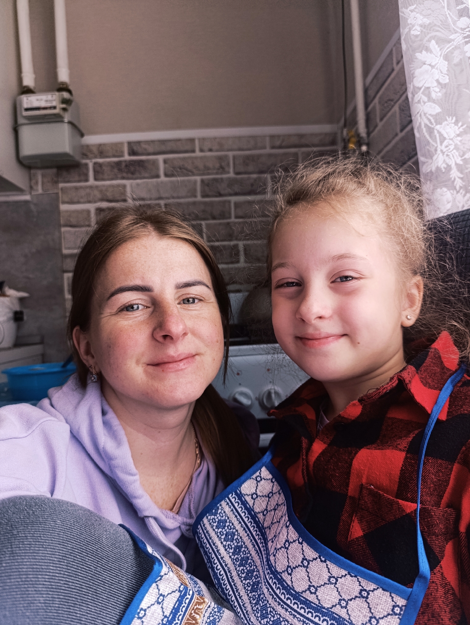 Two individuals, an adult female and a child, are centrally positioned in a close-up portrait. The woman, located on the left side of the frame, has light brown hair pulled back, revealing a face with visible freckles. She is wearing a lavender-colored hoodie and a gold necklace. The child, on the right, has light blonde hair styled in loose curls. She wears a red and black plaid shirt and a blue and white patterned apron.
The setting appears to be indoors, possibly a kitchen. Behind the subjects is a wall constructed of grey and white brick. To the upper left, a gas meter is affixed to the wall, and a white lace curtain is partially visible on the upper right. Further in the background, a white appliance, presumably a stove or oven, can be seen. A blue container and other items are discernible on a countertop in the background. The location is Unknown, Unknown.