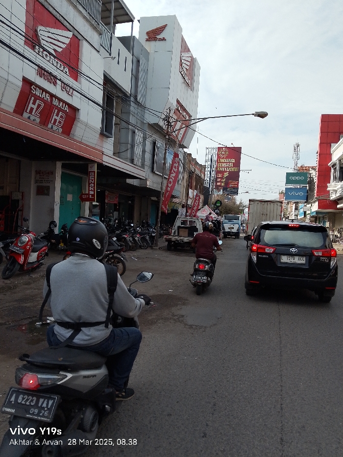 The image shows a street scene in Jatibarang, Indonesia, at coordinates -6.471 latitude and 108.302 longitude. The street is lined with buildings, including a Honda motorcycle dealership with a large red and white sign displaying the Honda logo. There are several motorcycles parked on the side of the street, and a man is driving a black scooter, which is directly in front of the camera. The man is wearing a gray shirt and black helmet, and he is looking straight ahead. Behind him is a row of parked scooters, and further up the street are several cars, including a black SUV that has stopped in traffic. In the background, there are more buildings, including a building with a large yellow sign with red text. There is a clear, blue sky in the background, and the street is paved with gray asphalt.