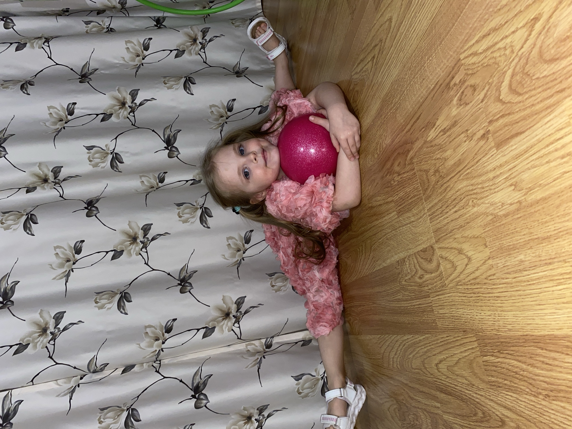 A young girl, likely of preschool age, with long light brown hair and blue eyes, is depicted lying on her back on a light-colored wooden laminate floor in Votkinsk, Russia. She is performing a straddle or split, with her right leg extended upwards towards a patterned curtain and her left leg extended along the floor. Dressed in a pink, ruffled outfit and white sandals, she holds a bright pink, sparkly ball against her chest with both hands. Her gaze is directed towards the camera, and a gentle smile suggests a playful and happy mood.
The indoor setting features a distinct light-colored wooden floor with visible grain patterns on the right side of the image. On the left, a patterned curtain hangs, adorned with a repeating design of light beige or white flowers and dark, thin stems with leaves against a light background. A fragment of a green object, possibly a hula hoop, is visible at the very top edge of the frame, near the curtain. The lighting indicates it is likely daytime indoors. There is no visible text in the image.