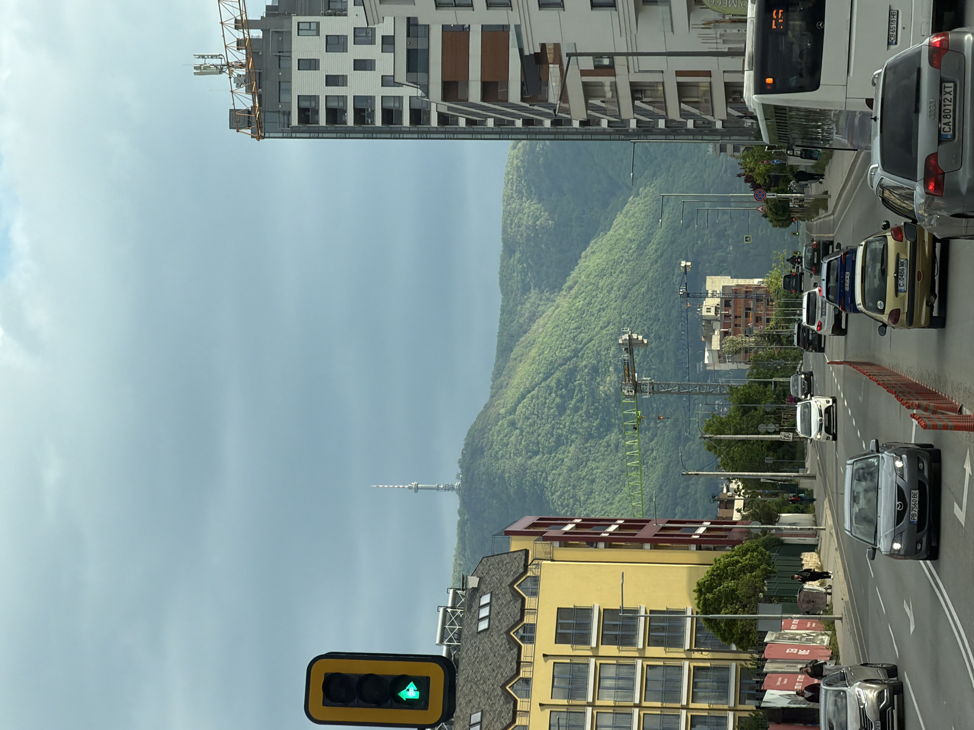 Here is a detailed description of the image:
The image captures a street scene in Sofia, Bulgaria, characterized by a mix of urban development and natural landscapes.
The most prominent natural feature is a densely forested mountain range visible in the background. A communications tower can be seen in the distance.
In the foreground, there is a busy street with multiple lanes of traffic. Cars are moving in both directions, and a large bus is visible among the vehicles.
The roadside has safety cones.
The scene is bordered by buildings on either side. There is a traffic light with a green arrow indicating that turns are permitted. The buildings vary in architectural style, ranging from modern high-rises to mid-rise structures with distinct design elements. At least two construction cranes suggest ongoing urban development.
People can be observed walking along the side of the street. The sky overhead is overcast, providing a soft, diffused light to the scene.