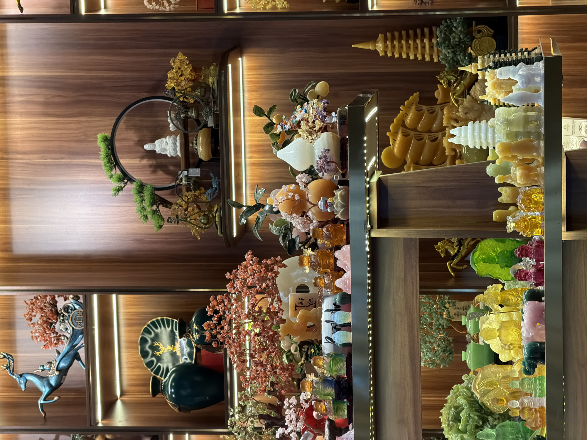 This image depicts a well-lit display shelf in what appears to be a shop or establishment in Bac Cam Ranh Ward, Vietnam. The shelves are made of wood and are illuminated by embedded strip lights. The shelves are densely packed with a variety of decorative objects, primarily made of glass or polished stone in various colors, including green, yellow, pink, orange, and white.
On the top shelf, a teal-colored deer sculpture is prominently displayed, pulling a chariot adorned with reddish-orange decorative stones. To its right, a dark wooden background is visible.
The second shelf from the top features two dark green vases, one larger than the other, and a decorative green plate with a gold emblem. There are also several small reddish-orange beaded decorative trees.
The third shelf holds a collection of small figures and decorative items. These include multiple small Buddha figures in different colors (green, orange, pink), a yellow deer-like sculpture, and a white gourd-shaped object. There are also small trees with colorful beaded foliage.
The bottom shelf showcases a wider array of colorful crystal or glass figures. These include more Buddha-like figures, elephants, a pink pig, and various other animal and abstract shapes. In the background of this shelf, a large, green, leafy sculpture is visible.
The overall impression is one of an ornate display of collectibles, possibly for sale, within a commercial setting in Bac Cam Ranh Ward, Vietnam. No people are visible in the image.