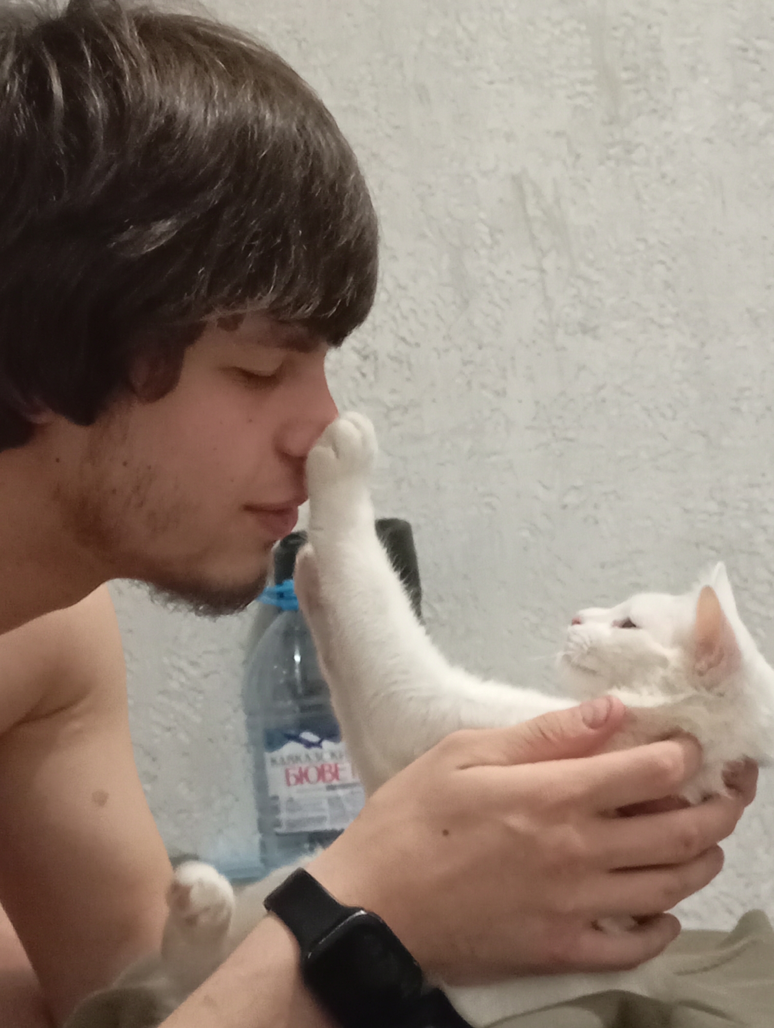 A fair-skinned man with dark hair and stubble on his face is shown in profile, right, with his eyes closed and the tip of his nose touching the paw of a white cat. The cat, which is held by the man’s right hand, has its left paw raised to the man’s face. A water bottle labeled "KASKAD BIOVET" is behind the man and cat. The bottle is partially obscured. The man is wearing a black smartwatch on his left wrist. The scene is likely indoors and against a white wall. Based on the bottle's label, the image was likely taken in or near Krasnodar, Russia.