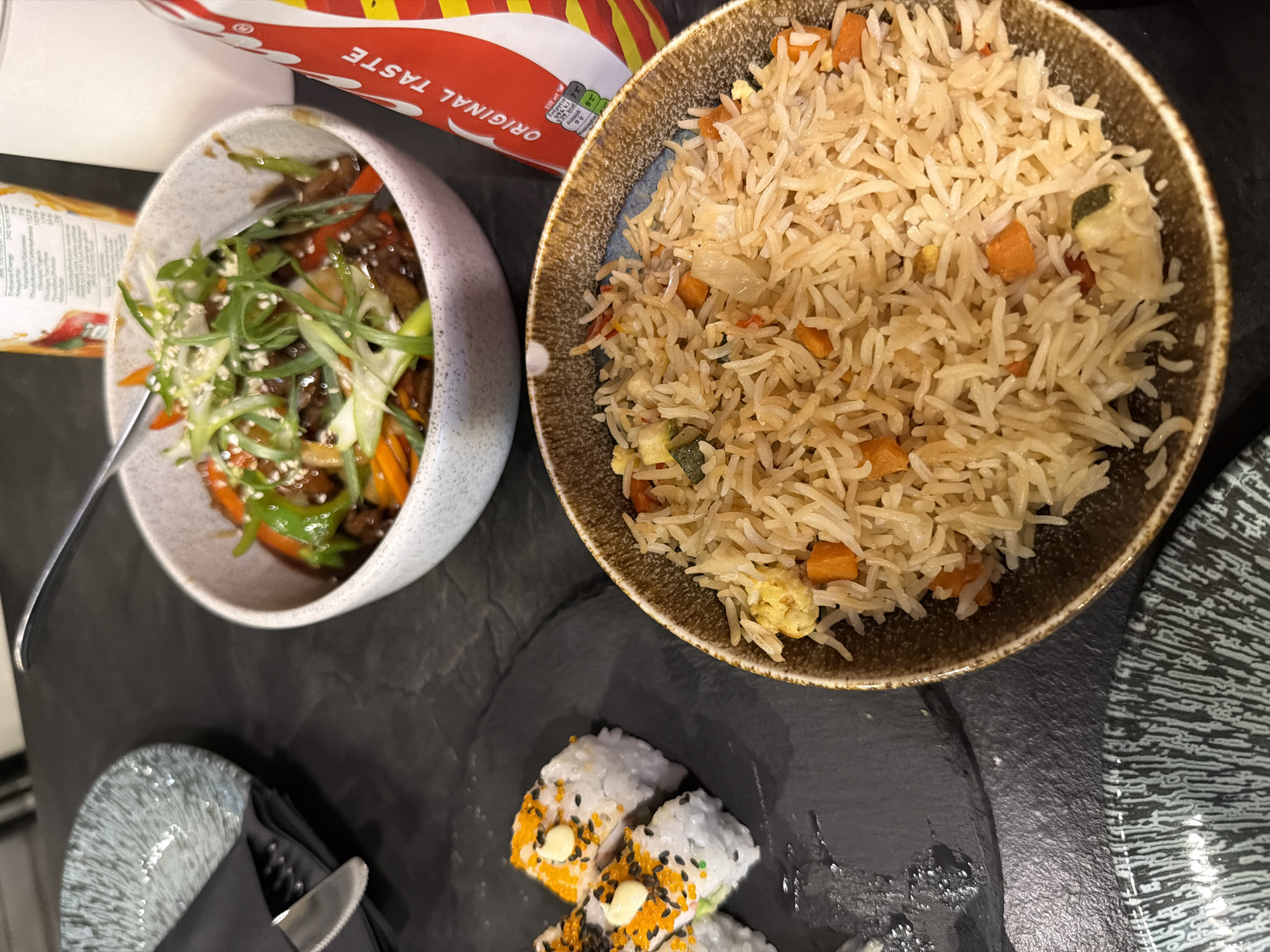 The image shows a close-up view of a table at a restaurant in Kiti, Cyprus, featuring a meal. In the foreground, a bowl of fried rice with diced carrots and zucchini is prominently displayed. To the left, a plate holds several pieces of sushi rolls, topped with white sauce and orange roe. Behind the sushi, a dark slate placemat is visible, with a decorative plate and cutlery to the left. In the mid-ground, a bowl contains a stir-fry dish with beef, green peppers, carrots, and scallions, sprinkled with sesame seeds. A fork rests in this bowl. To the right, a partially visible bottle of Coca-Cola, with its iconic red and white branding and "ORIGINAL TASTE" text, stands next to a glass of a clear liquid. The overall setting suggests a casual dining experience.