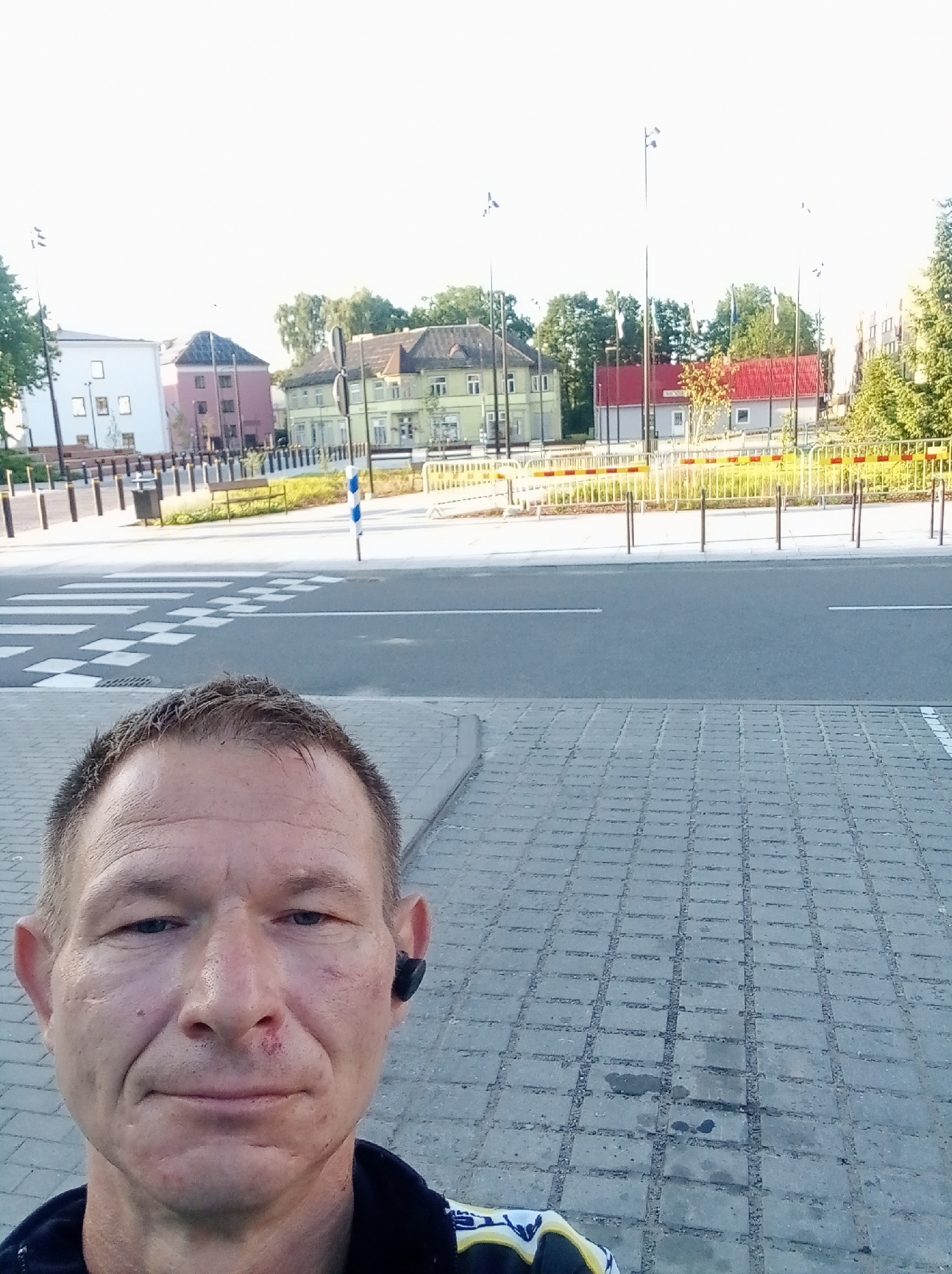 The image is a first-person perspective selfie taken in the city of Tapa, Estonia. The foreground shows the head and shoulders of a man with short, graying hair and a weathered complexion. He is wearing a dark jacket with yellow and blue accents and has a black wireless earbud in his right ear. There is a small scrape on his left cheek and a minor abrasion on the bridge of his nose. The background reveals a street scene with a pedestrian crossing marked with white stripes and checkered squares. Beyond the street, there are several buildings of varying architectural styles, including residential and commercial structures, interspersed with trees and lampposts. Yellow and red striped barriers are visible along the sidewalk. The sky is bright and overcast, suggesting daytime. The man appears to be standing on a paved area with a patterned brick surface.