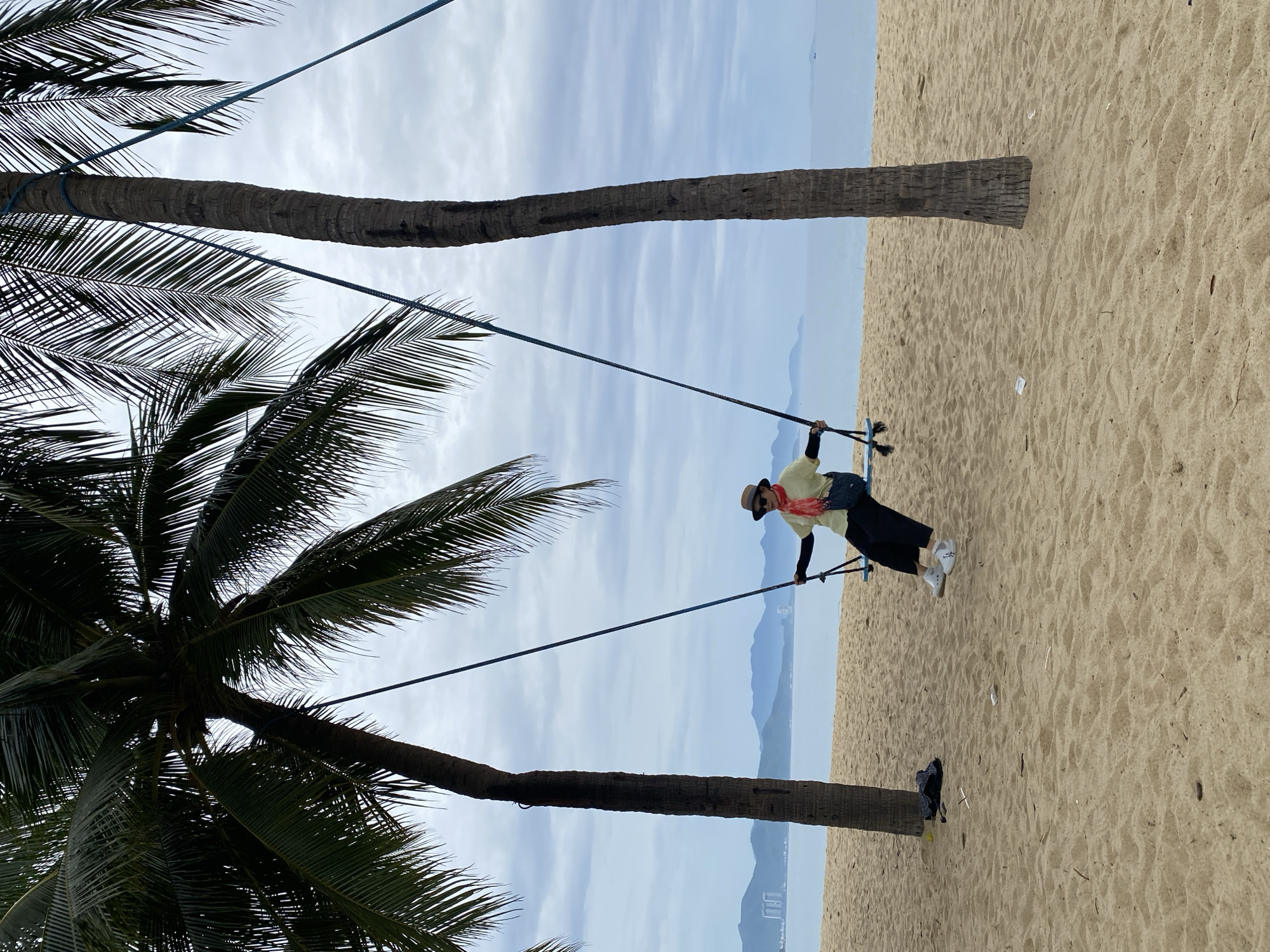 A woman is seated on a swing between two palm trees on a sandy beach in Nha Trang, Vietnam. She is wearing a yellow shirt, black pants, white sneakers, a hat, and a red scarf. The swing is suspended by blue ropes attached to the palm trees. In the background, the ocean is visible, with distant mountains and buildings along the coastline under a cloudy sky. A black bag is on the sand near the left palm tree.