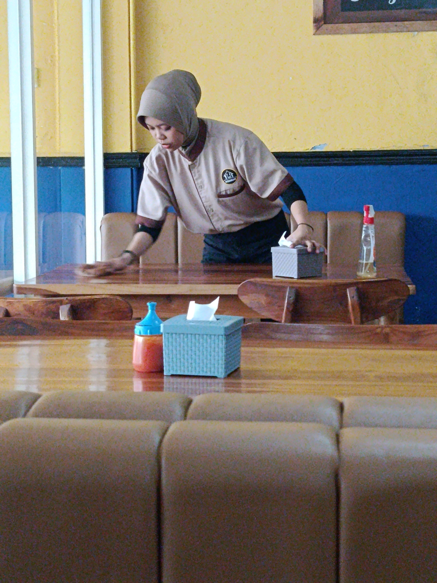 The image depicts a restaurant interior in Bekasi, Indonesia. A young woman, dressed in a light brown uniform and a headscarf, is wiping down a wooden table with her right hand. She is positioned behind a row of plush, tan-colored seats and in front of a blue wall and a yellow wall. On the table, in front of her, are a teal tissue box, a small orange bottle with a blue cap, and a spray bottle with a red cap. The tables and chairs appear to be made of wood. The overall ambiance suggests a casual dining establishment.