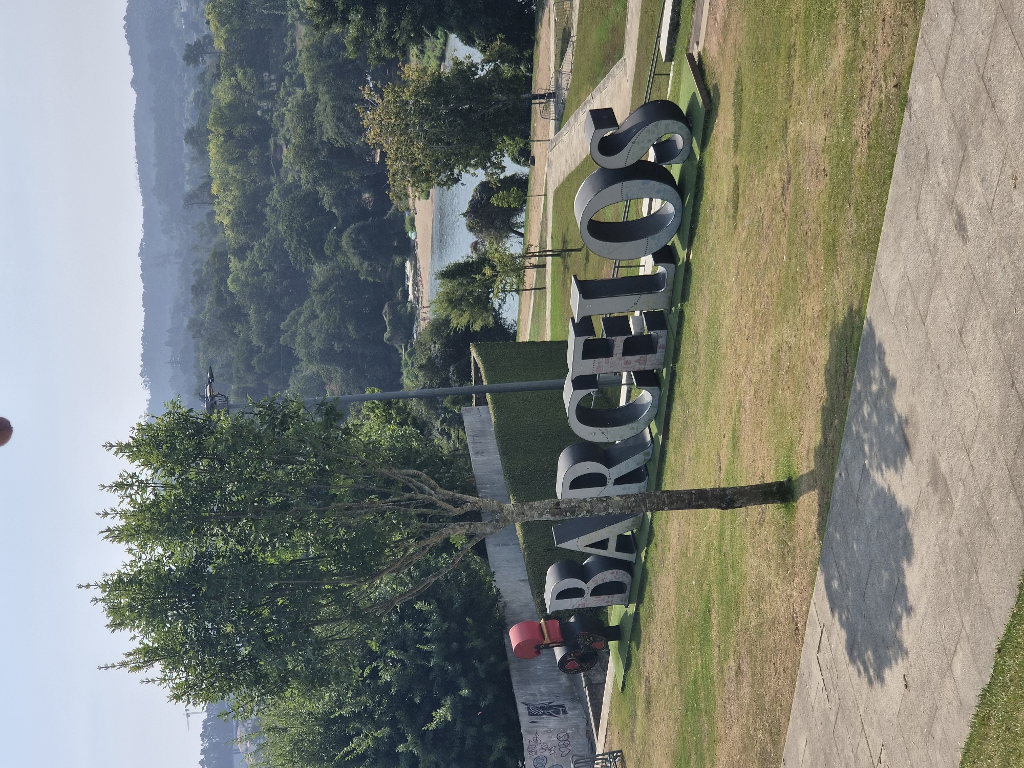 A wide-angle, outdoor, daytime shot captures a large, sculptural sign spelling out "BARCELOS" in bold, dark gray 3D letters on a vibrant green lawn. To the left of the sign, a vintage-style red and black movie camera sculpture is visible. A slender tree stands in front of the sign, casting a shadow on a paved walkway in the foreground. In the background, a serene river with a sandy beach and a few distant figures can be seen, bordered by lush green trees and rolling hills under a bright, hazy sky.