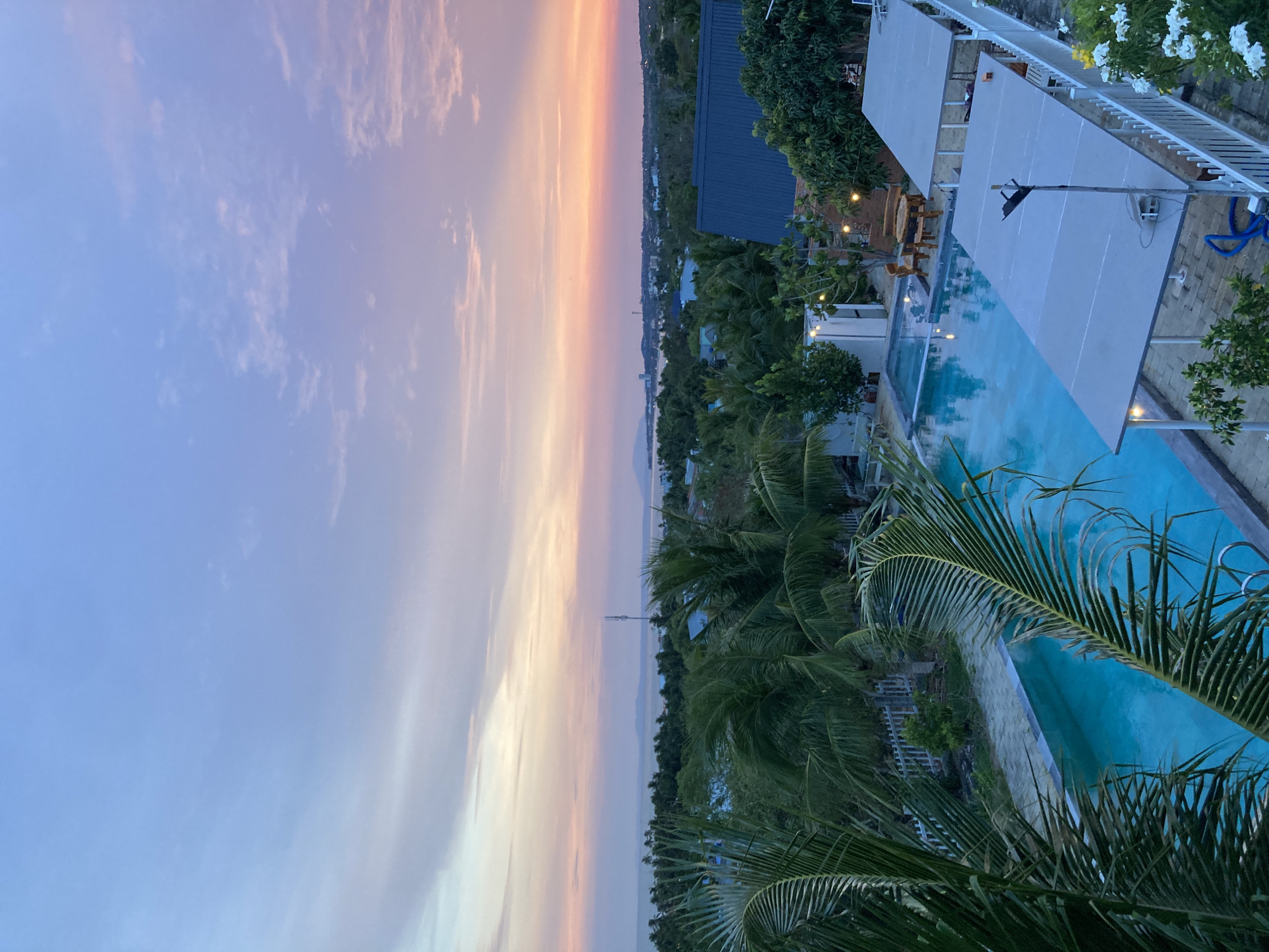 The image captures a view overlooking a swimming pool and a distant coastline at sunset. The scene is set in what appears to be a tropical resort or private residence in Mũi Né, Vietnam. The foreground is dominated by the green fronds of palm trees, partially obscuring the pool area. The pool's turquoise water contrasts with the pale gray canopies shading a lounge area next to it. Beyond the trees, a cityscape stretches along the coast, fading into the soft hues of the sunset sky, with colors ranging from pale blue to orange and pink. A tall structure, possibly a communication tower, is visible in the distance on the coast.