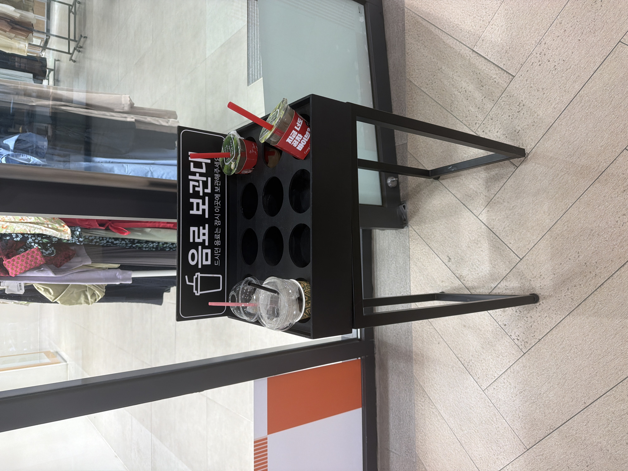 The image shows a black metal stand with circular slots for holding drinks, situated near a storefront. The stand has three drinks in it: two plastic cups with straws, and one cup with "Gong Cha" branding. Korean text is printed on the stand, which appears to be a drink storage facility. The environment suggests an indoor location, possibly a store, with clothing visible through a glass partition and patterned floor tiles. The photo was taken in Gimpo-si, South Korea.