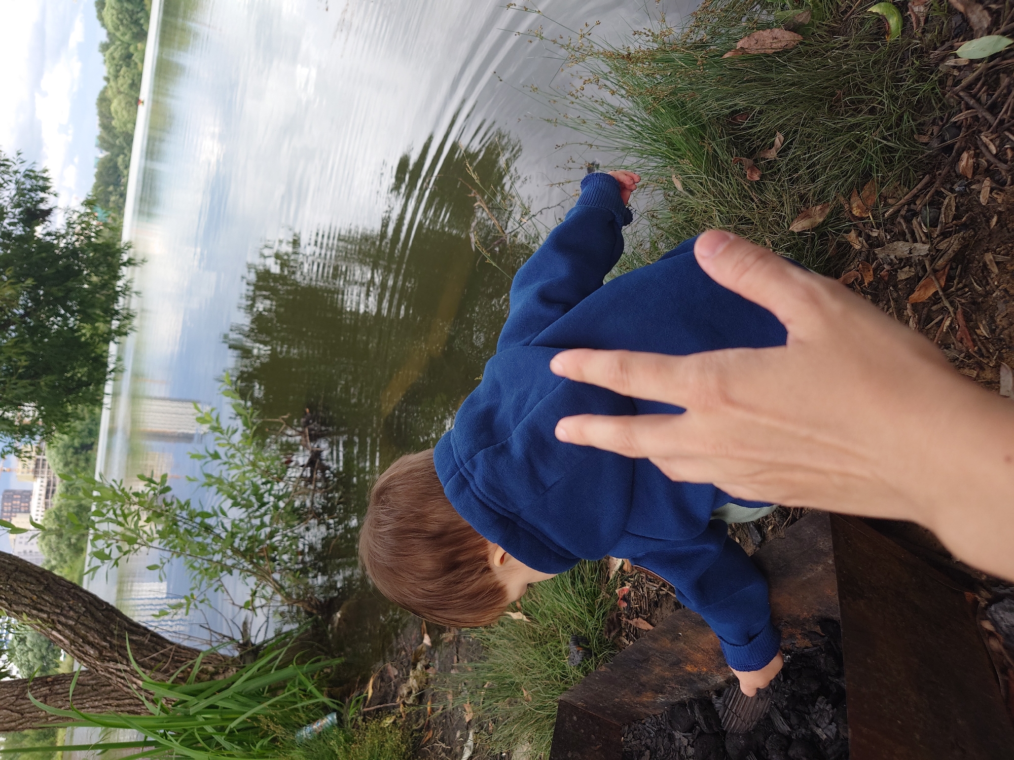 An overhead, close-up shot captures a young child, seen from behind, wearing a blue hooded jacket and light-colored pants. The child is crouched down, appearing to interact with a metal fire pit filled with dark coals. Next to the child, the water's edge of a lake is visible, with tall green grass growing along the bank. In the background, the hazy outlines of buildings suggest an urban or suburban setting, with trees and foliage framing the body of water. The foreground is dominated by a hand, with fingers extended, partially obscuring the child and the fire pit. The scene appears to be set in the city of Kommunarka, Russia.