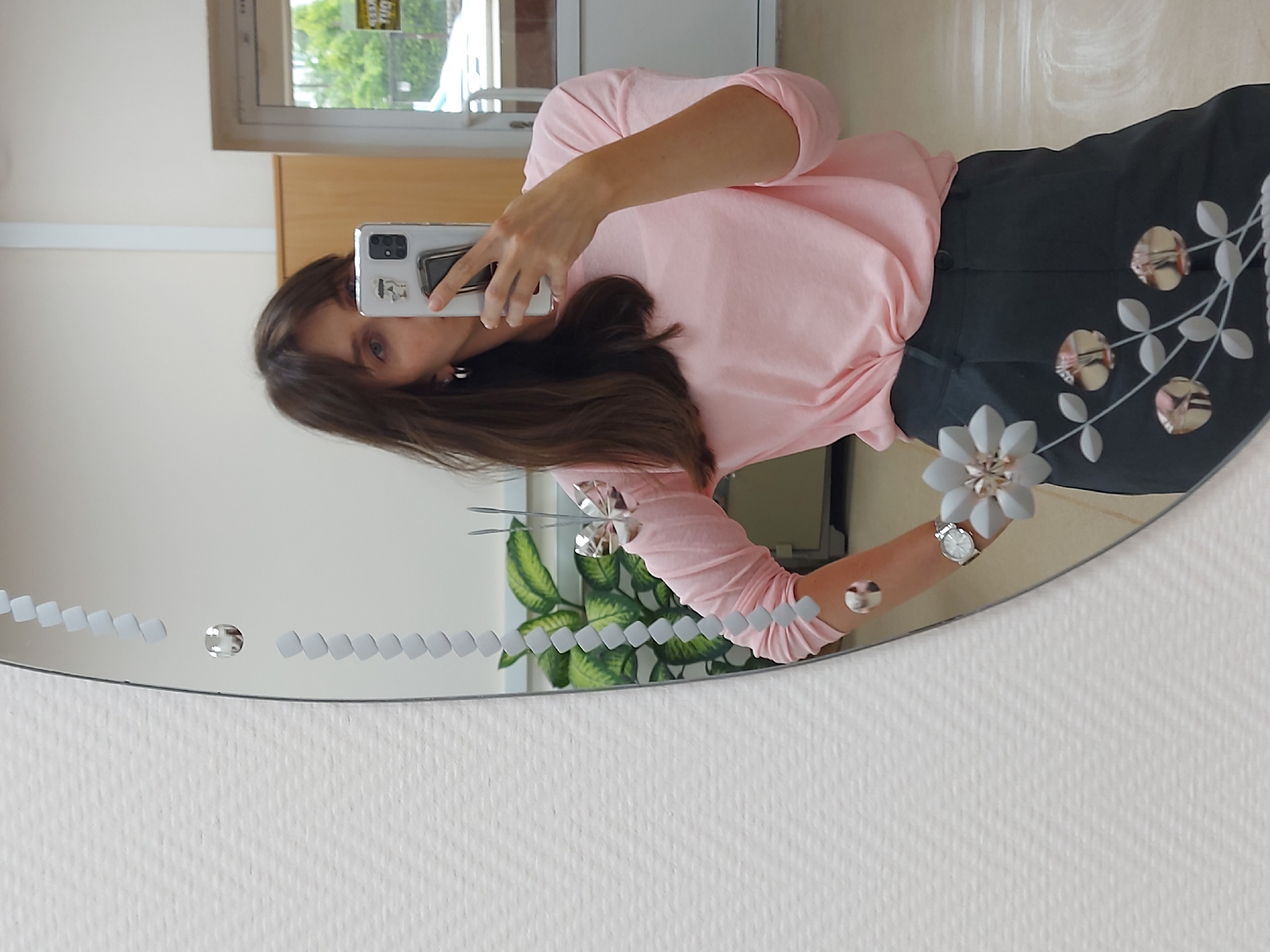 A brunette woman is taking a selfie in a mirror. She's wearing a pink long-sleeved shirt and dark pants or a skirt with floral embellishments. She holds a white smartphone up to her face. The mirror is round and has decorative etched patterns around the edge. Reflected in the mirror is a plant and what appears to be part of an interior space with a window and beige cabinets or walls. The wall behind the mirror has a textured white surface. Based on previous data, the location is likely Chaikovsky, Russia.