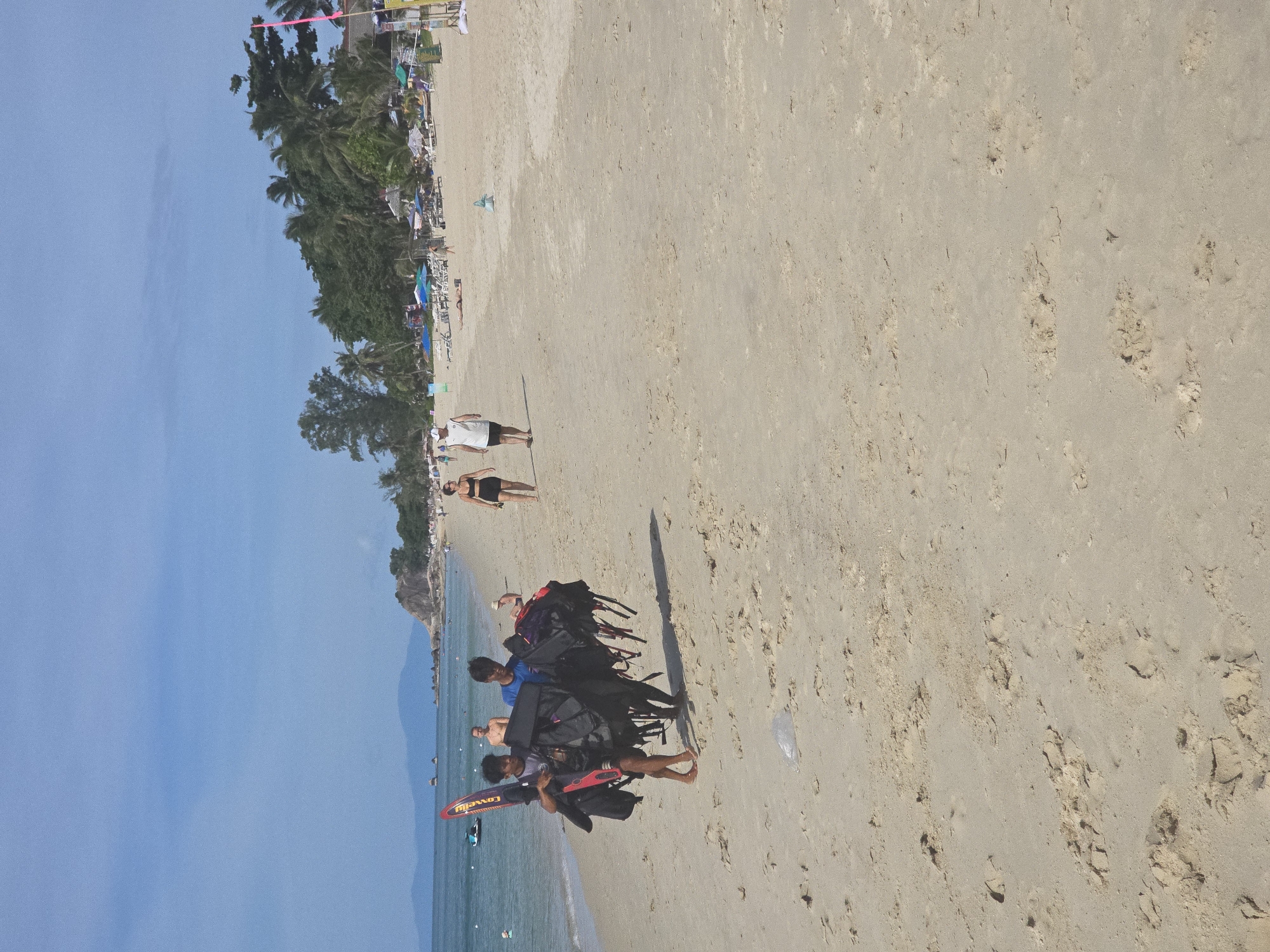 Two young men are walking on a sandy beach in Baan Lamai, Thailand, carrying a collection of black bags and a red and yellow surfboard. The men are wearing casual clothing. The beach is lined with palm trees and foliage. In the distance, there are mountains and the ocean. The sky is clear and blue. Several beach chairs and umbrellas are visible further down the beach, suggesting a populated area.