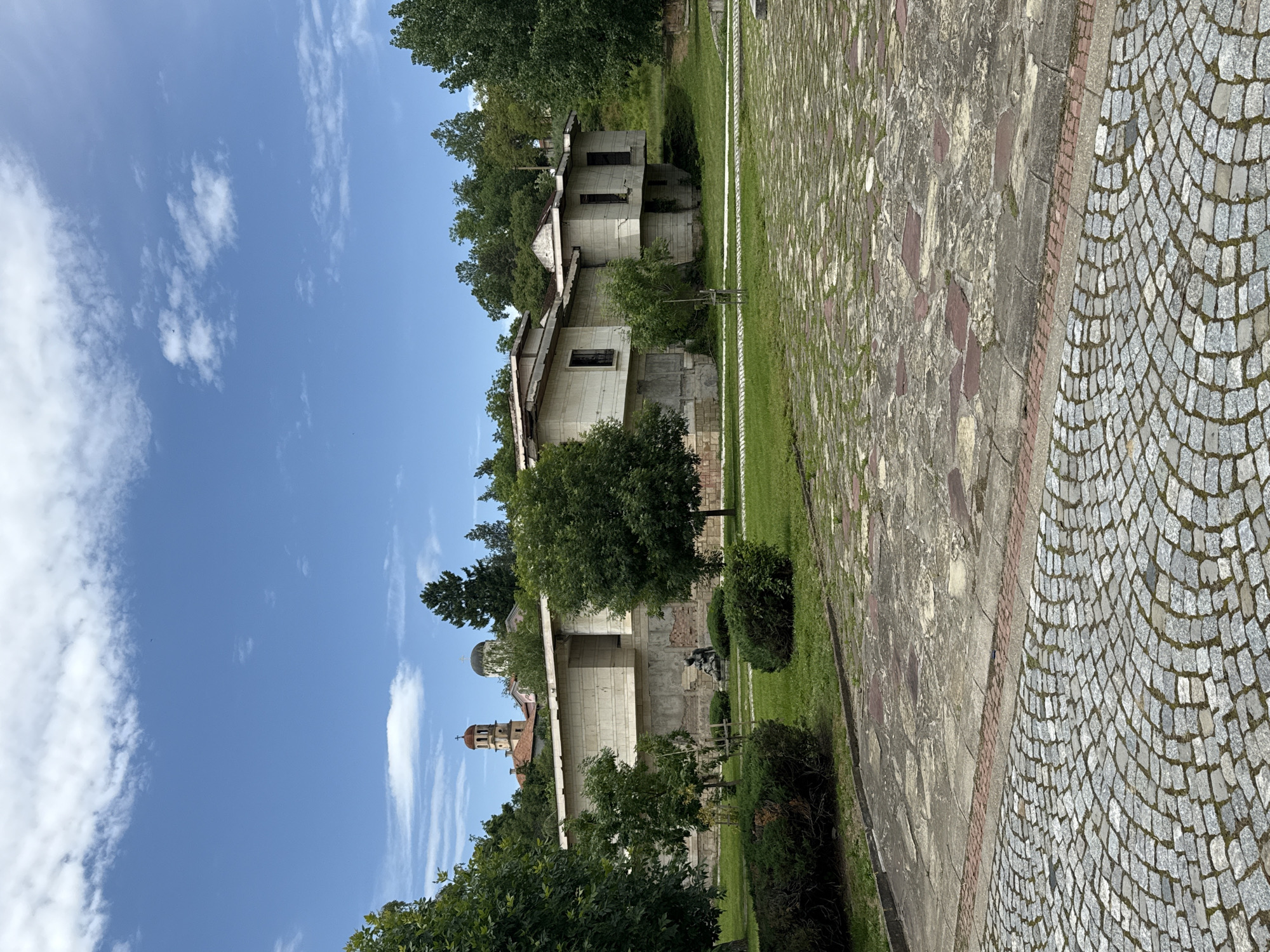 The image shows an exterior scene in Strazhitsa, Bulgaria, featuring a landscape with buildings and paved areas. In the foreground, there is a walkway made of small, gray cobblestones, adjacent to a wider paved area with larger, irregular stones. Green grass grows between the paving stones.
Behind the paved areas, there are trees and bushes that partially obscure a set of light-colored, two-story buildings. The buildings appear to be unfinished or undergoing renovation, with visible concrete walls and missing windows. Behind these buildings, a church tower with a red roof is visible. The sky is blue with scattered white clouds.