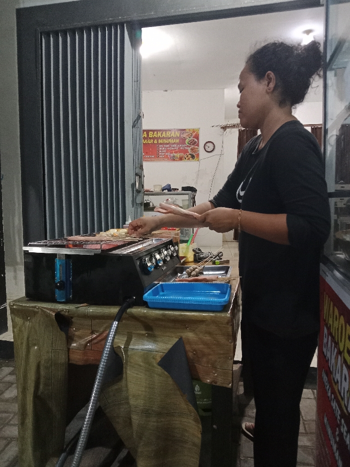 The image presents a nighttime outdoor grilling operation in Turunsih, Indonesia. A female individual, positioned in profile facing right, with dark hair secured in a bun and wearing a dark long-sleeved top, is actively engaged in grilling. Her left hand is manipulating items on a black gas grill equipped with four front-facing control knobs. The grill rack holds several skewers with various food items, including sausages and other meats. Adjacent to the grill on a counter, a blue rectangular plastic tray and a metal tray contain additional skewers with uncooked items, notably dark, spherical objects resembling meatballs. A flexible metallic hose extends from the left side of the grill downwards. The counter surface is draped with a dark olive-green fabric. Behind the subject, an open doorway reveals a brightly illuminated interior space. A red and yellow banner on the interior wall advertises "A BAKARAN" (grilled items) and "BAKAR & MINUMAN" (grilled items & drinks), listing food options such as "ANEKA SATE." A circular wall clock and a white refrigerator are visible within the indoor space. To the left of the doorway, a dark grey accordion-style security gate is partially open. A red and white display case, partially visible on the far right, shows text including "WAROE" and "BAKARAN." The ground consists of light-colored paving tiles.