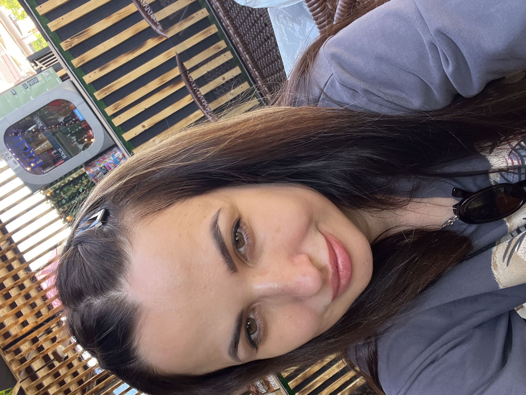 A young woman with long, dark brown hair is taking a selfie outdoors. She is wearing a gray t-shirt and a silver necklace with sunglasses hanging from it. Her hair is partially pulled back with a silver clip. She has brown eyes and is smiling slightly. The background shows a wooden fence and a cooler filled with drinks. The lighting suggests it is daytime.