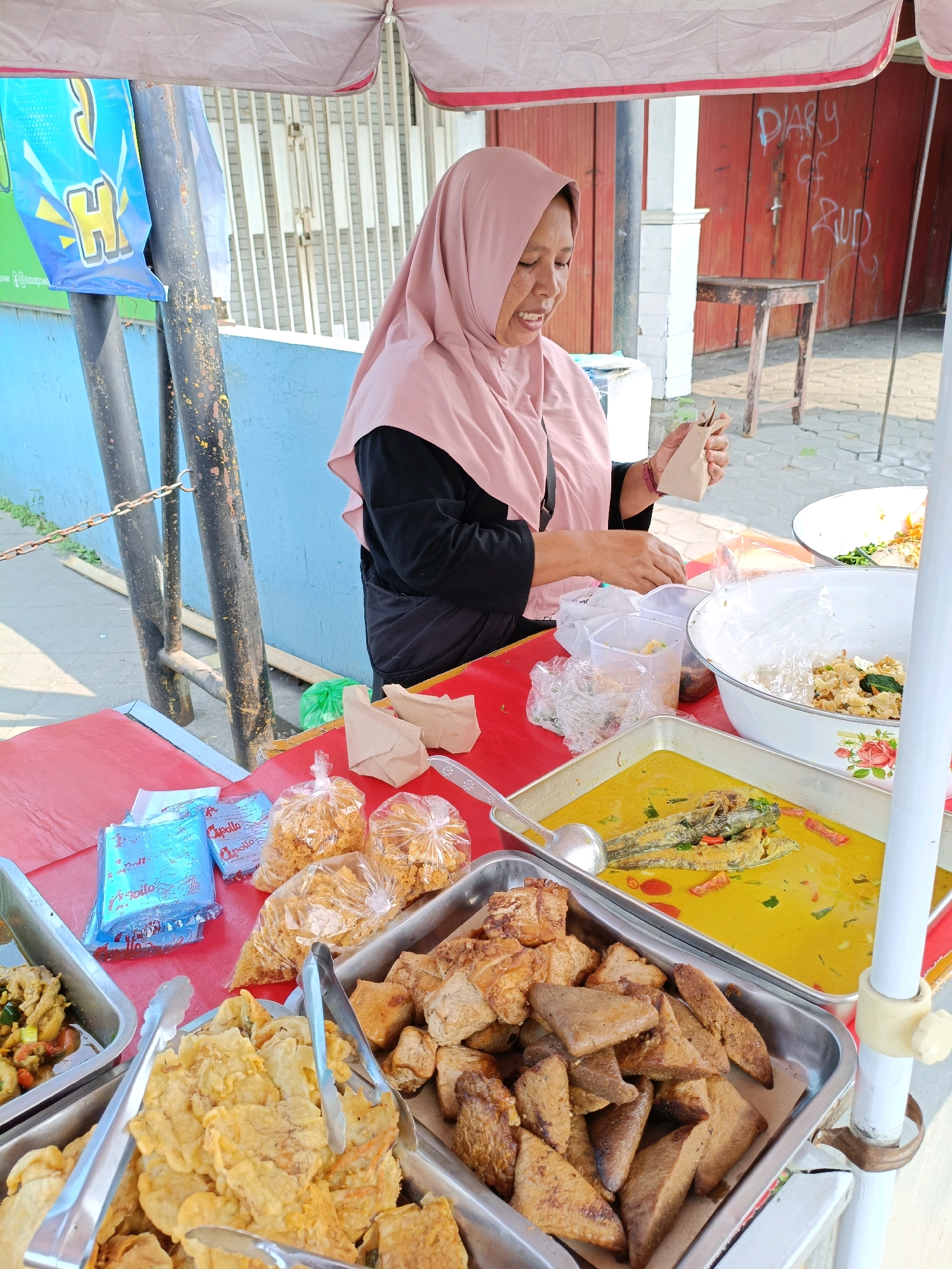 An outdoor food stall is set up under a canopy with a pink border, likely in Maguwoharjo, Indonesia. A woman wearing a pink hijab and a black shirt is behind the stall, attending to customers. She is holding a small brown paper bag and appears to be in the process of packaging food. The stall features various food items displayed in metal trays. In the foreground, there are trays filled with fried tempeh and other fried items, as well as a tray of fish in a yellow curry. In the background, there are bags of snacks, blue packages, and a large bowl of food. The stall is positioned in front of a building with red doors and graffiti that reads "DIARY OF RUD." A metal pole with chains is visible on the left side of the image, and a blue banner with yellow lettering hangs from it. The ground is paved with interlocking bricks, and there is a wooden table and stool behind the stall. The overall scene depicts a bustling street food vendor with a variety of Indonesian dishes.