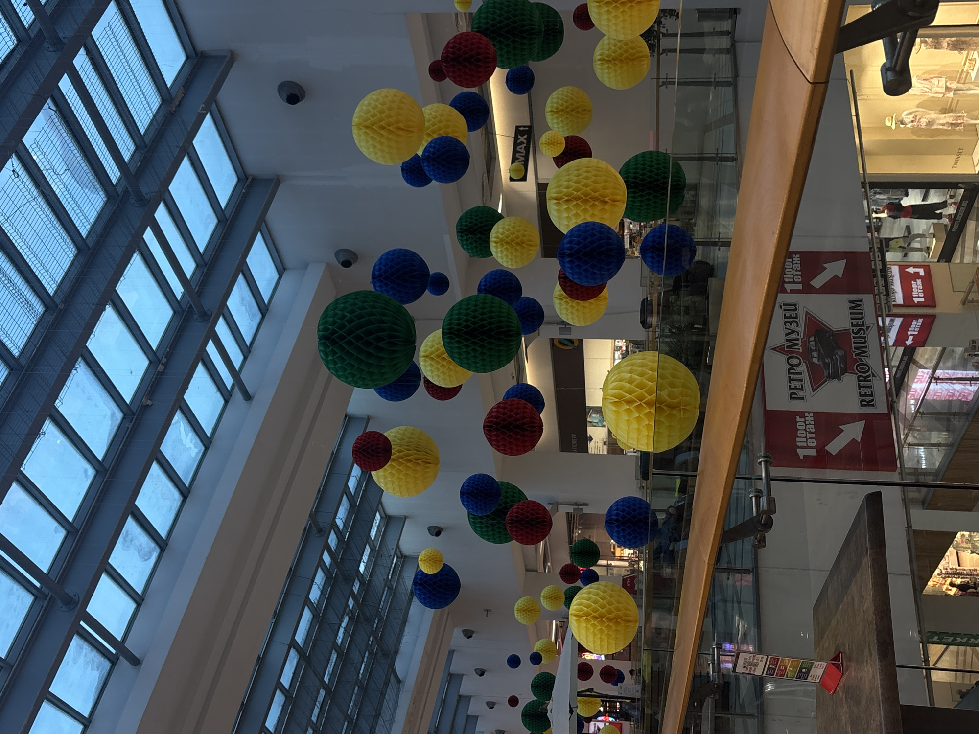 The image depicts an interior view of what appears to be a shopping mall in Varna, Bulgaria. Above, a ceiling composed of numerous rectangular windows framed by a grid of metal bars is visible, letting in natural light. Suspended from the ceiling are numerous colorful, honeycomb-shaped spheres in yellow, blue, green, and red, arranged in a decorative pattern. On the right side of the image, one can see the edge of a balcony with displays from various shops, including signs in both English and Bulgarian, such as "RETRO MUSEUM" and brand names. The "MAX1" sign and multiple storefronts indicate a bustling commercial environment. The scene is well-lit, indicating daytime, and the perspective suggests the photograph was taken from a lower level, looking up through multiple stories of the shopping center.