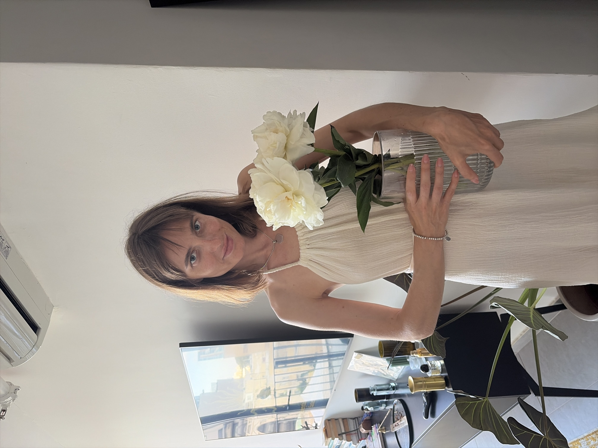 A woman with shoulder-length brown hair and green eyes is holding a clear, ribbed glass vase filled with white peonies. She is wearing a cream-colored, sleeveless dress with a halter neck and a silver necklace. The woman is looking towards the camera with a slight smile. Her right arm is extended, holding the vase, while her left hand rests on the vase as well. She is wearing a delicate silver bracelet with a small heart-shaped charm on her left wrist. The background appears to be an indoor setting with a white wall and a glimpse of a television screen showing an outdoor scene. To the right, there are houseplants and some decorative items on a table. The lighting suggests it might be daytime, with natural light coming from the side. The overall mood of the image is calm and serene.