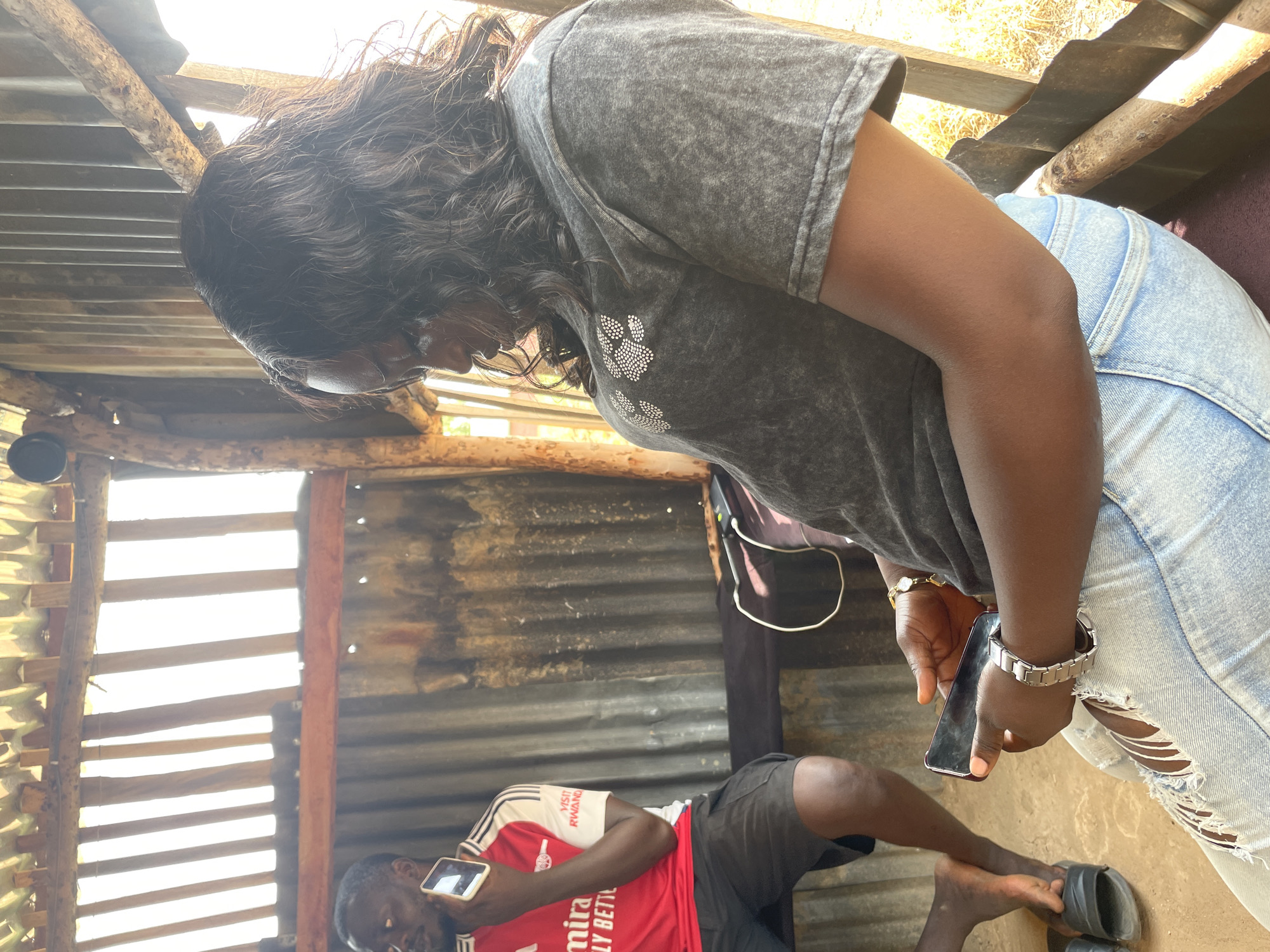 A medium shot captures two individuals indoors, with the primary subject, a woman with dark, wavy hair, seated in the foreground. She is wearing a distressed denim outfit and a gray t-shirt adorned with a white paw print design. Her hands are engaged with a smartphone, and she wears a silver watch and a gold bracelet. To her left, a man in a red and white soccer jersey and black shorts is visible. He holds a white smartphone to his ear. The environment appears to be a rustic, open-air structure with corrugated metal walls and a thatched roof, suggesting a rural or semi-developed setting in Durumi, Nigeria. The lighting indicates it is daytime, with sunlight filtering through the roof. A dark bag and a charging cable are present on a surface between the two individuals.