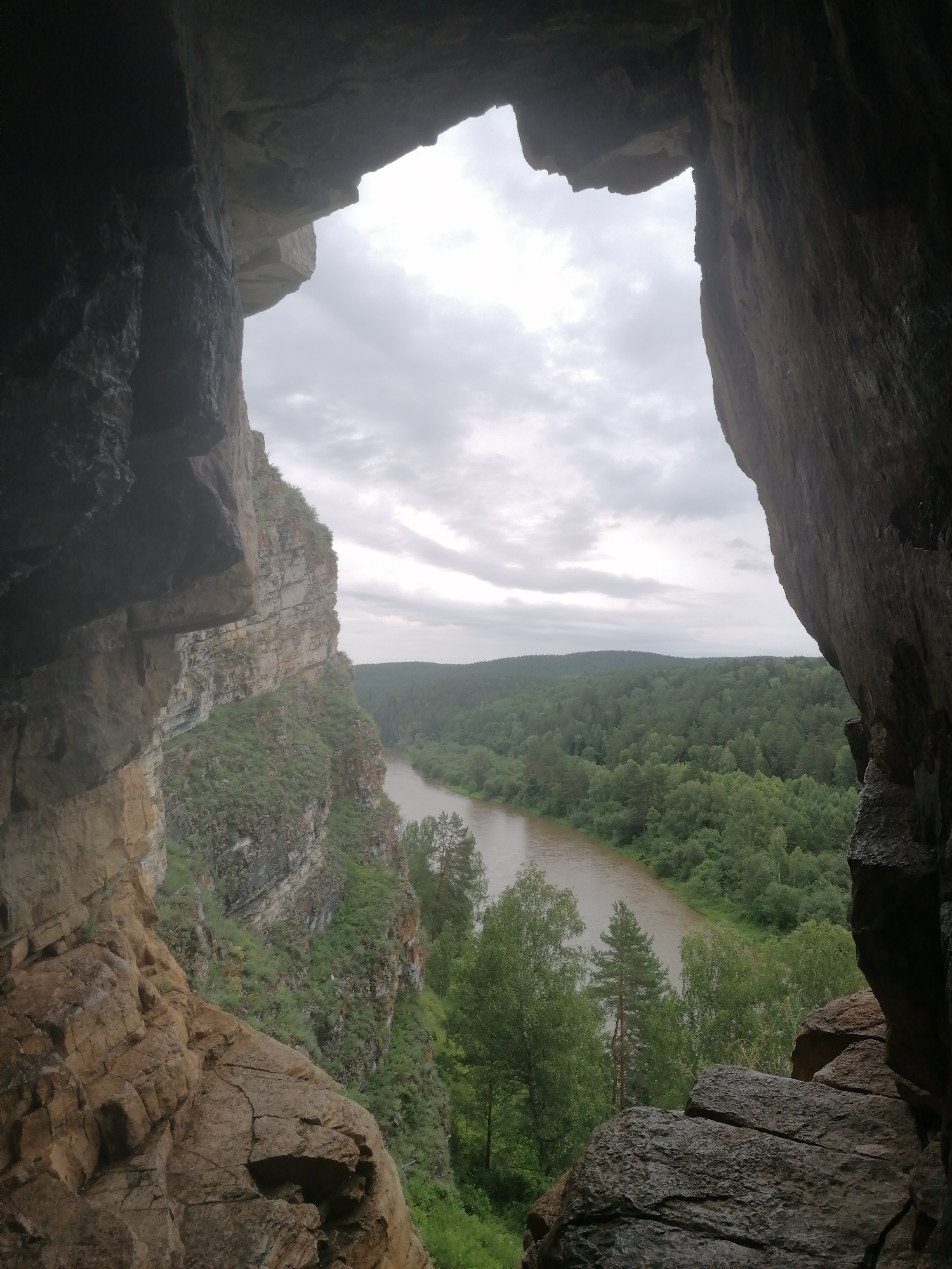 This media file depicts a view from inside a cave or rock overhang, looking out at a scenic natural landscape. The foreground is dominated by the rough, textured rock formations that form the mouth of the cave. These rocks are a mix of browns and grays, with visible cracks and variations in their surface.
Through the opening, a wide, muddy river flows through a valley. The river is bordered by lush, green forests that cover rolling hills. The trees are dense and appear to be a mix of deciduous and coniferous types. The sky is overcast with gray clouds, suggesting a cloudy or possibly rainy day. The overall lighting is subdued.
The scene appears to be a natural, undeveloped location, likely a natural park or wilderness area. The perspective suggests the viewer is standing within a geological formation, possibly a cave or a natural arch, observing the expansive view. There is no text visible in the image. The mood conveyed is one of natural grandeur and perhaps a sense of awe or peace inspired by the vastness of the landscape. The time of day appears to be daytime, but the overcast sky makes it difficult to determine a precise hour. The location is identified as Lagerevo, Russia.