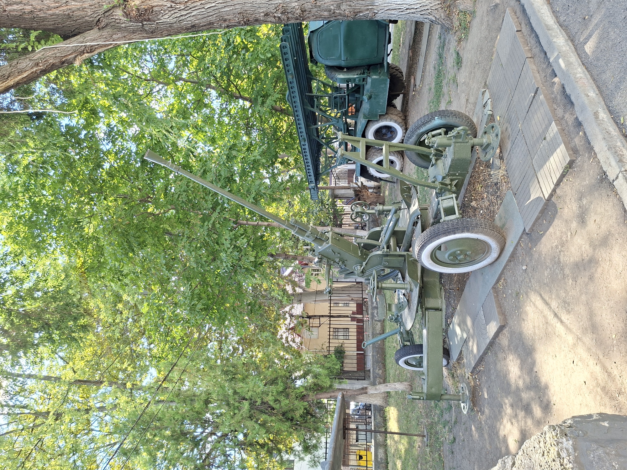 In the city of Chișinău, Moldova, a military exhibit is set up outdoors. The primary focus is a green, towed anti-aircraft gun positioned on a wooden platform. To the right of the gun, a large green military truck with a rocket launcher mounted on its back is visible. The environment is characterized by lush green trees providing shade, and a clear blue sky. In the background, a building with a yellow facade and a fence can be seen. The ground is a mix of dirt and gravel, with some dry leaves scattered around. There are no people present in the image. The scene appears to be a static display, likely in a park or memorial area.