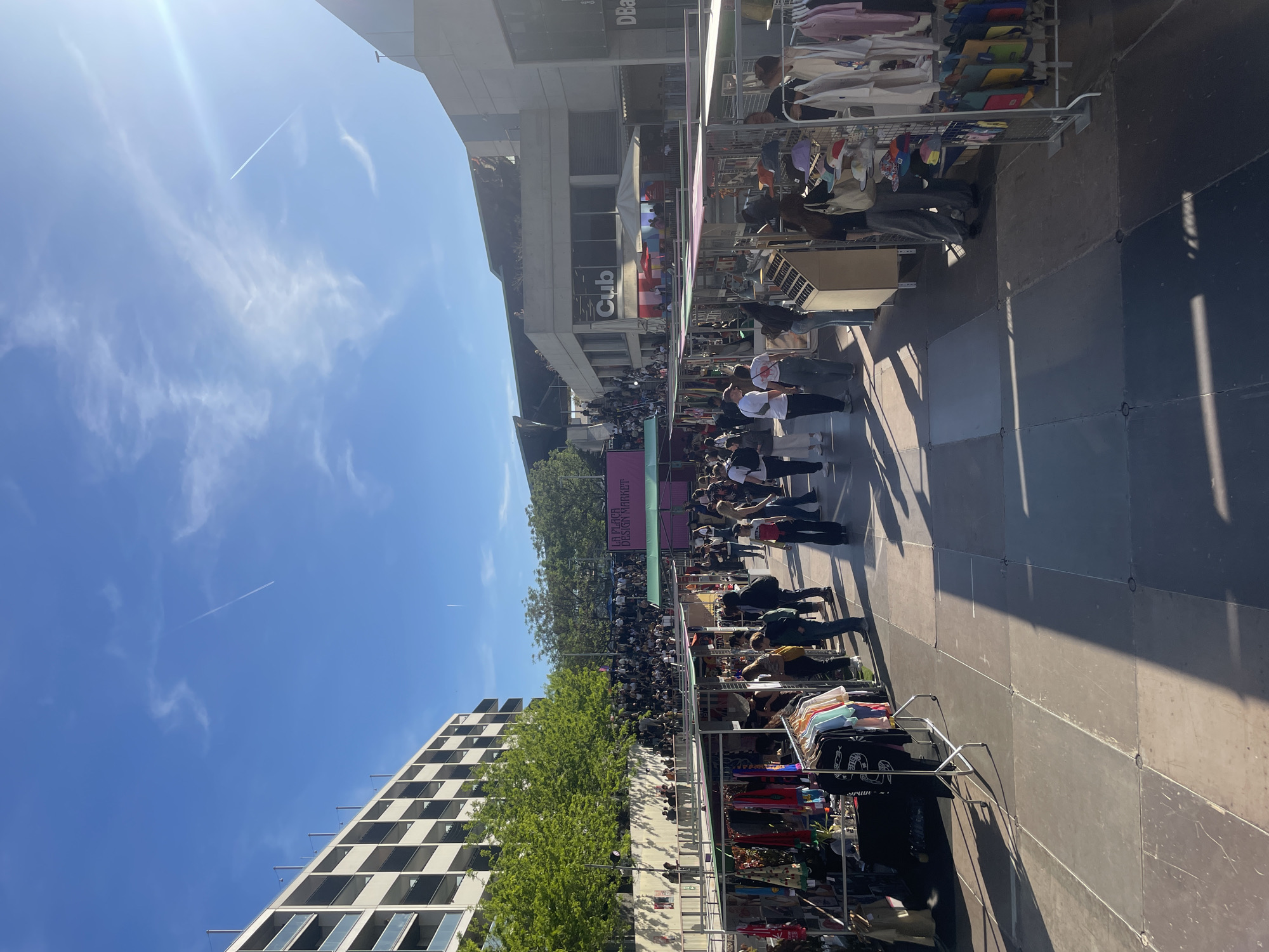 The image captures an outdoor market scene in Barcelona, Spain. A crowd of people is observed amidst vendor stalls displaying clothing and other goods. Buildings surround the market area. The sky is blue with contrails visible. The scene is well-lit, suggesting a sunny day.