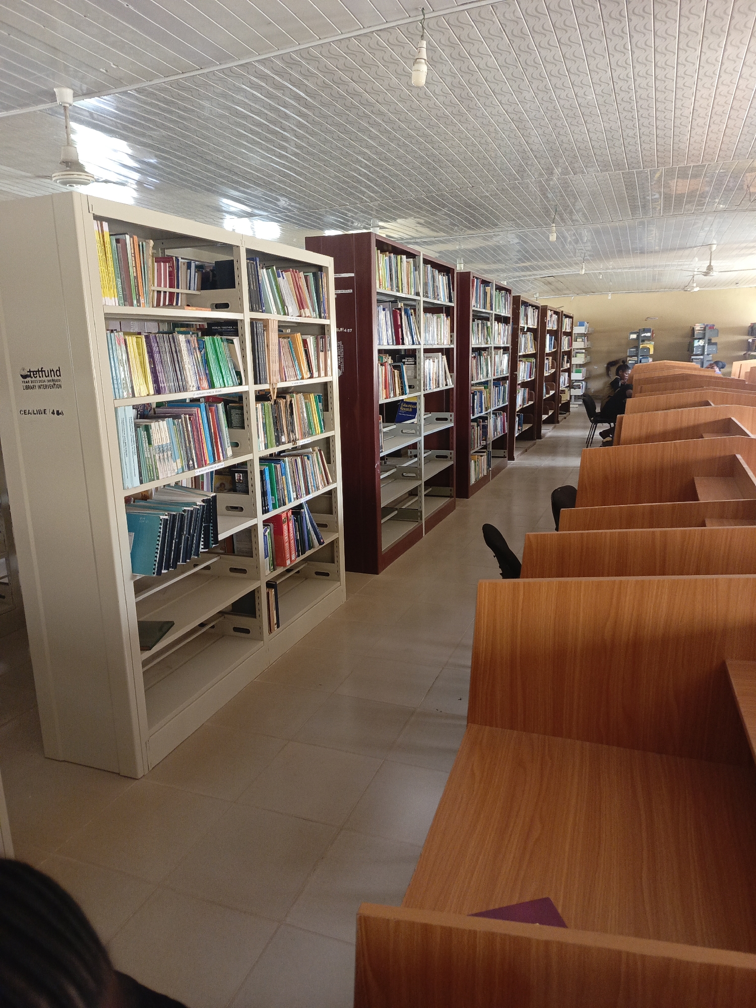 The image depicts the interior of a library in Akwanga, Nigeria. Rows of bookshelves are filled with books, and wooden carrels provide individual study spaces. A person can be seen seated at a desk in the distance. The library has a tiled floor and a ceiling with fluorescent lights. The TETFund logo on one of the bookshelves indicates the library's intervention.