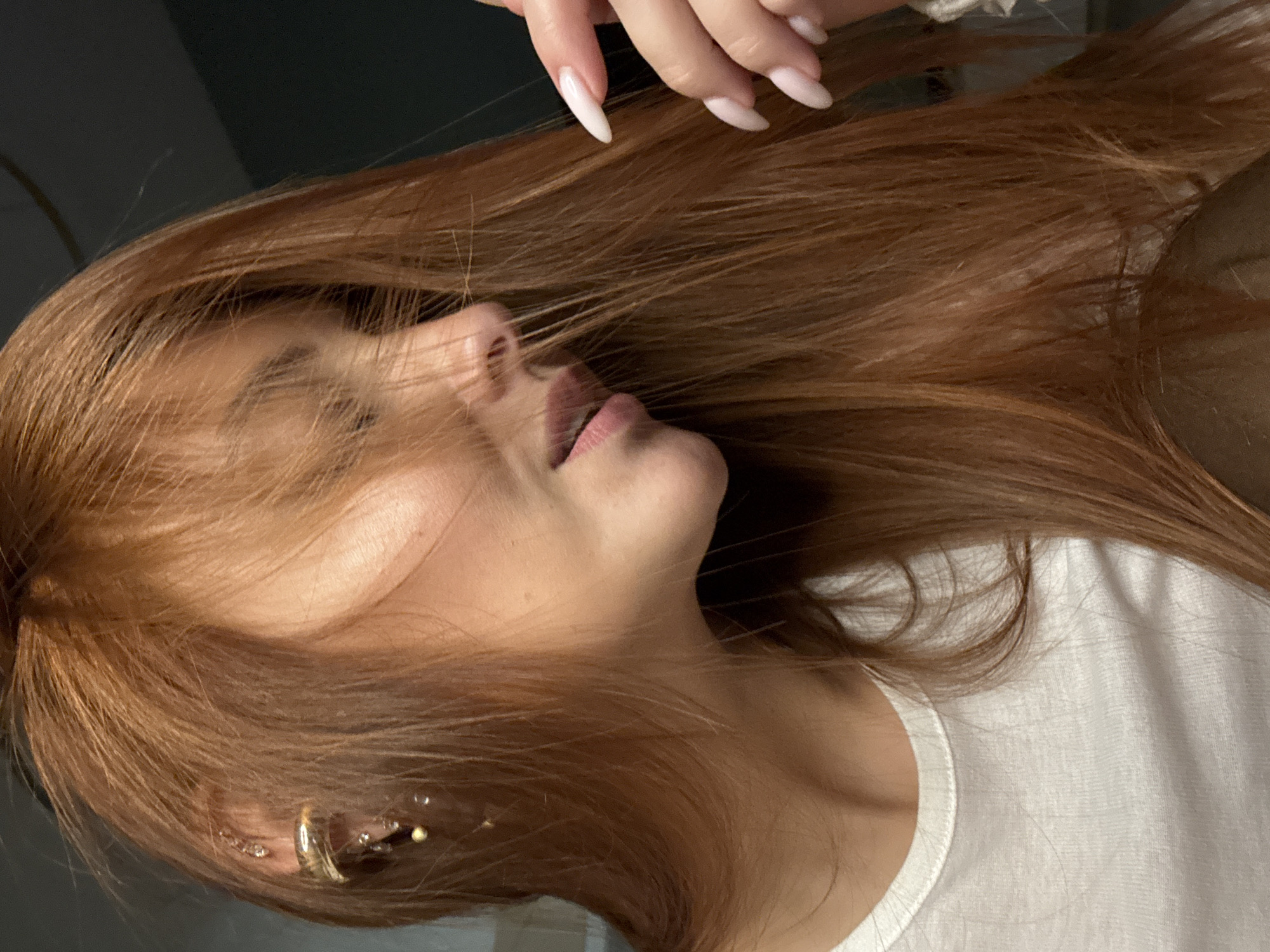 This close-up portrait, captured in Chaikovsky, Russia, features a person with long, flowing reddish-brown hair. The image is composed vertically, showing the individual's profile from the nose down, with their eyes and upper face largely obscured by their voluminous hair. A hand with long, light-colored, pointed nails is visible in the upper part of the frame, appearing to gently touch or arrange the hair.
The person is wearing a simple white t-shirt, its neckline and shoulder visible in the lower right. Their mouth is slightly open, suggesting a relaxed or contemplative expression. The ear is adorned with several pieces of jewelry, including a prominent gold-toned hoop earring and multiple smaller, delicate studs with clear stones. The background is dark and indistinct, providing no specific location cues beyond the given context, and suggesting an indoor setting or low-light conditions. The lighting highlights the texture and color of the hair. No visible text is present in the image. The overall impression is one of quiet intimacy and casual elegance.