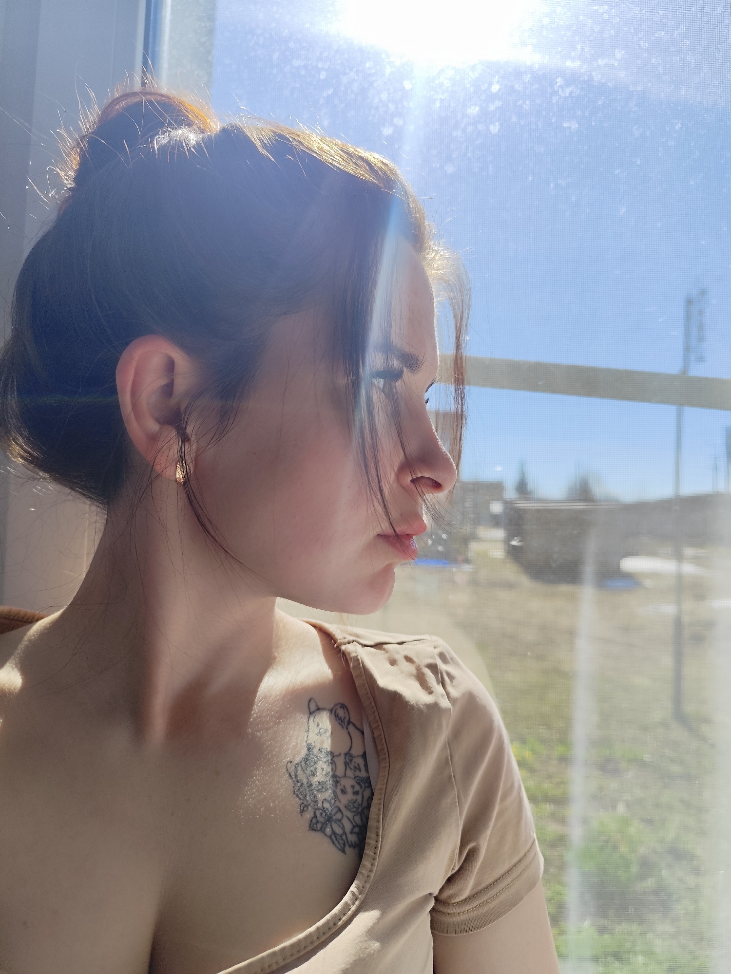 A fair-skinned woman with dark hair pulled into a bun stands indoors, gazing out a window on a sunny day. She has a small gold earring and a tattoo on her upper chest of what appears to be stylized cats. The window offers a blurred view of buildings and what looks like vegetation. Based on the city name, the location is Фоки, Russia.