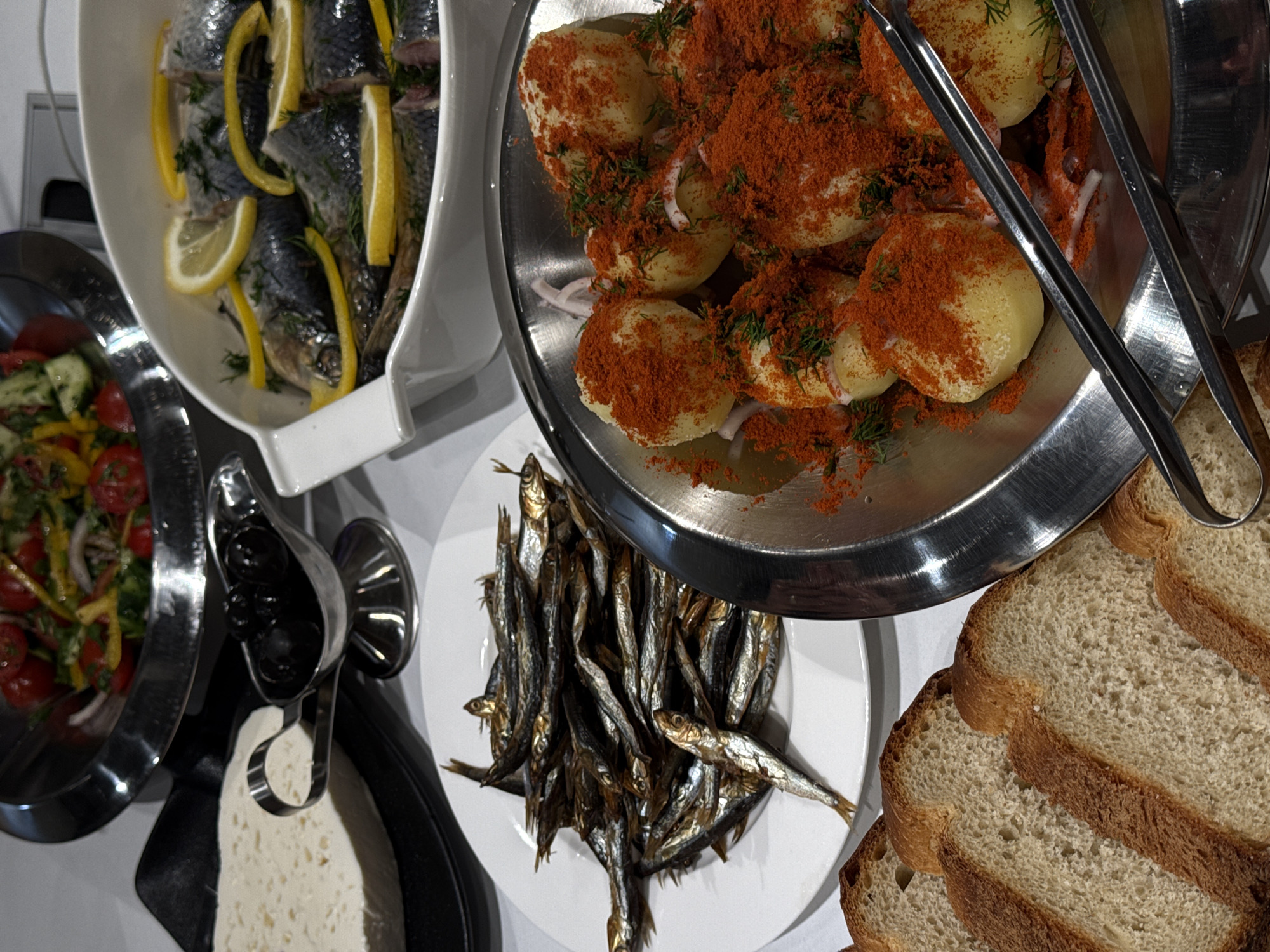 A high-angle shot shows a buffet spread. From left to right are a bowl of salad, a white dish of sliced herring with lemon and dill, a black serving spoon holding olives, a dish of white cheese, a white plate piled with small fried fish, a stainless steel bowl of boiled potatoes covered in paprika, a pair of serving tongs, and stacked slices of bread. The location appears to be an indoor dining setting, possibly in Yerevan, Armenia, as suggested by the presence of herring, a traditional Baltic dish common in Yerevan restaurants, and the style of the buffet. No people are visible in the frame.