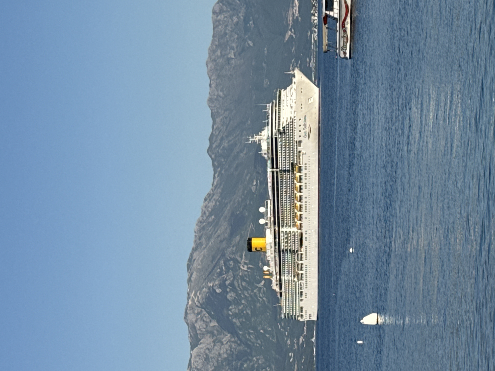 A photograph shows a large, white cruise ship docked in the harbor of Đenovići, Montenegro. The ship, positioned vertically, has a yellow smokestack with a prominent "C" symbol. It appears to be a Costa Cruises vessel, likely the Costa Deliziosa based on the name appearing on the side. Numerous windows line the ship's exterior. Behind the ship, a rugged mountain range rises steeply. The sky is a clear, light blue. The water in the harbor is a deep blue, with visible reflections and a smaller boat nearby.