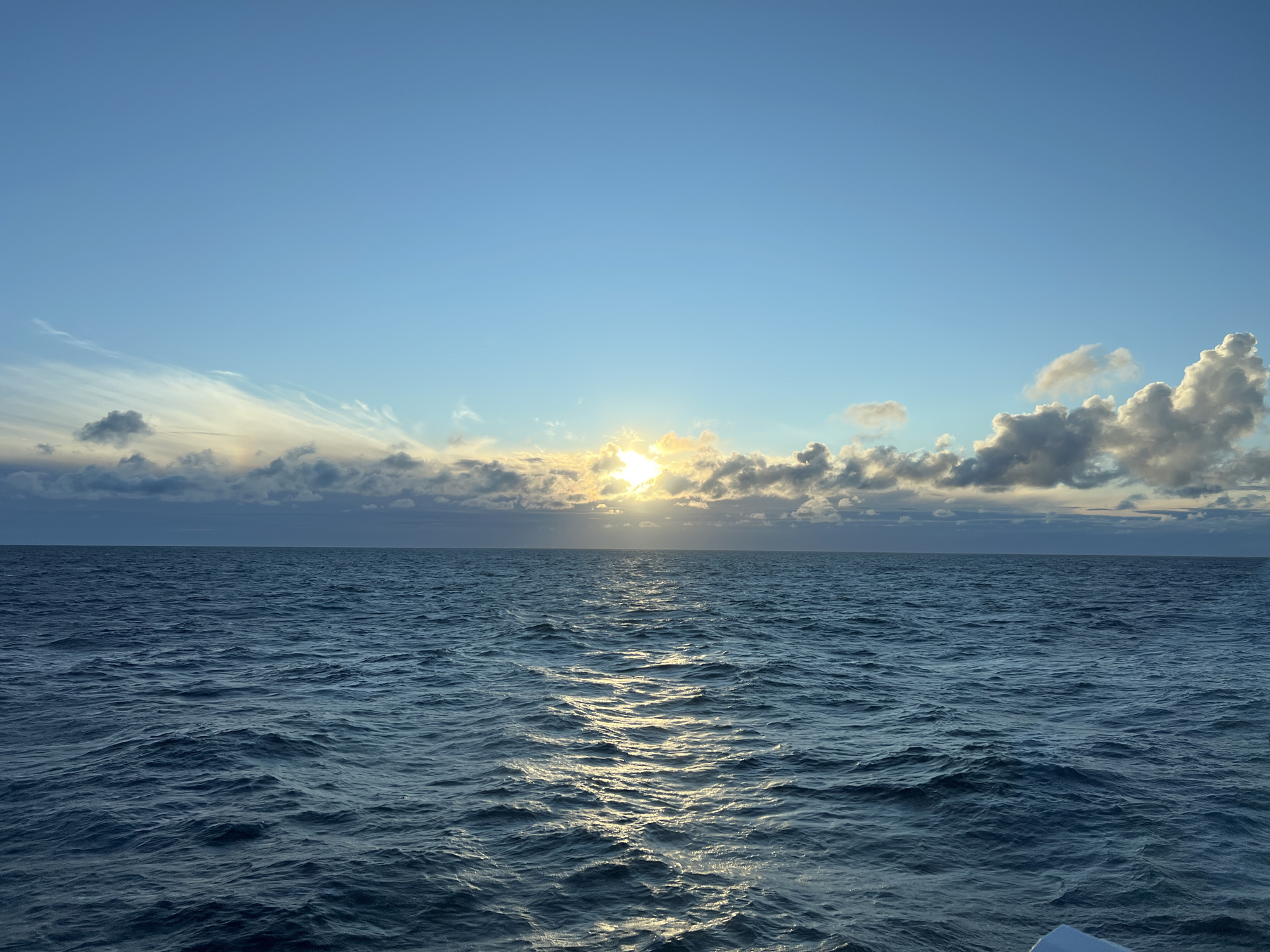 The image shows a vast expanse of the ocean under a clear blue sky, with the sun partially obscured by scattered clouds. The sun's rays create a shimmering path on the water's surface. The water is deep blue with moderate waves. The horizon line is clearly defined where the ocean meets the sky. The scene appears to be a tranquil seascape, possibly during sunrise or sunset. There is no indication of the city of Unknown, Poland, or any specific landmarks within the image.