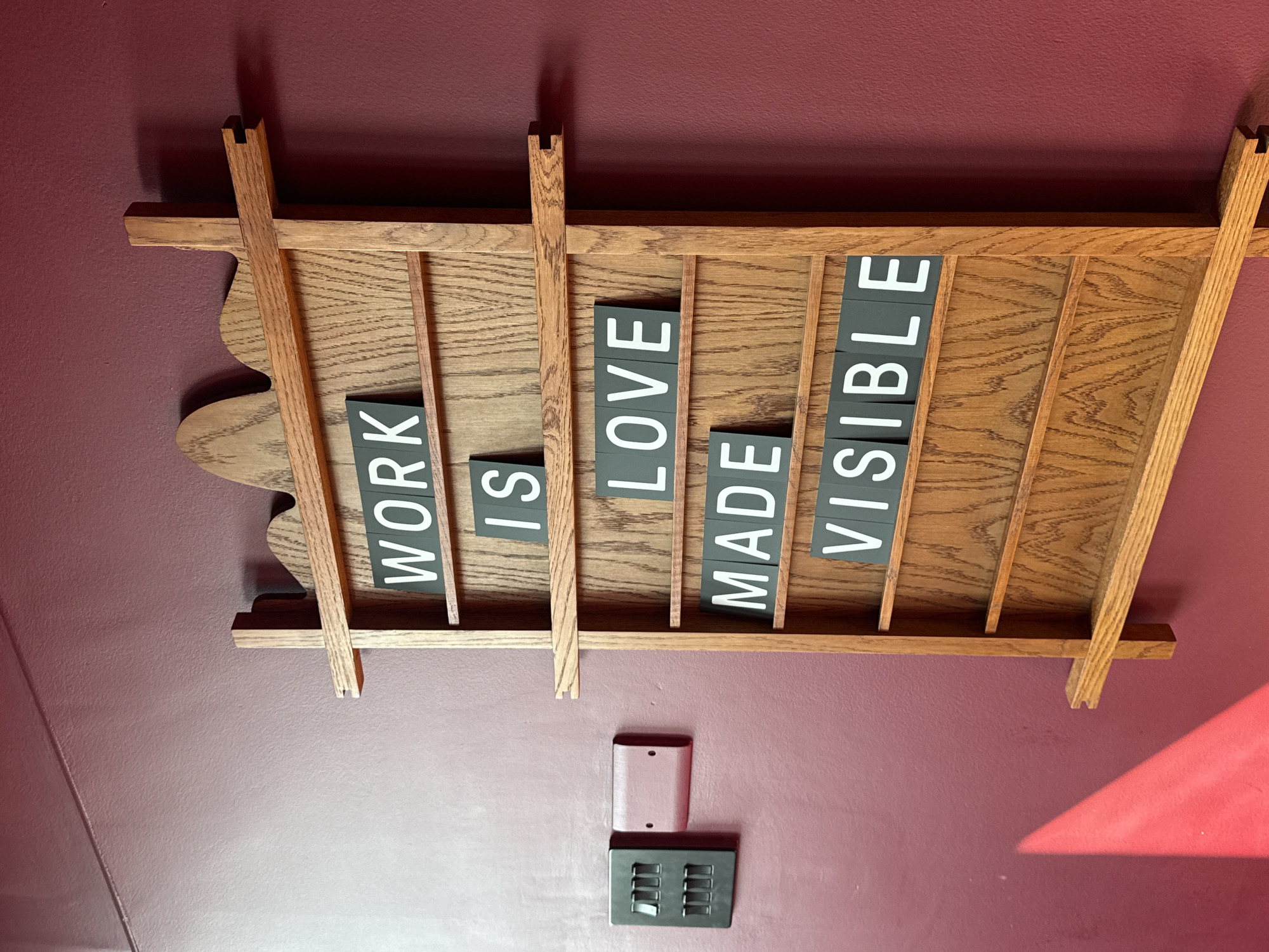 The scene depicts an interior wall, presumed to be in the city of London, United Kingdom. A vertical, light brown wooden display rack, featuring a decorative scalloped top edge and multiple horizontal grooves, is centrally mounted on a matte maroon-colored wall. Within these grooves, dark grey square tiles with white capital letters spell out the phrase: "WORK IS LOVE MADE VISIBLE," arranged over five lines. To the left of the wooden rack, a black electrical switch plate containing six individual rocker switches is mounted on the wall. Immediately to the right of this switch plate, a smaller, flush-mounted, rectangular metal plate, painted to match the maroon wall color and secured by two visible screws, is present. A prominent, triangular patch of bright reddish-orange light illuminates the lower left section of the wall. No individuals are visible.