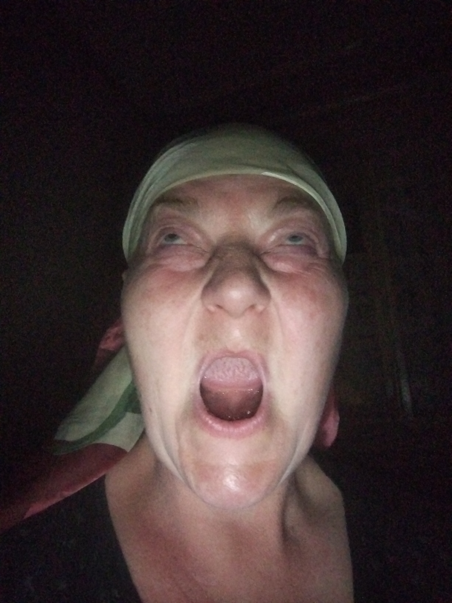 A close-up, wide-angle, low-light selfie shows a middle-aged woman with her eyes rolled back and mouth wide open, as if screaming or yawning. She is wearing a light-colored headscarf and a dark shirt. The background is dark and appears to be indoors. There is no information to suggest the image was taken in Horishni Plavni, Ukraine.