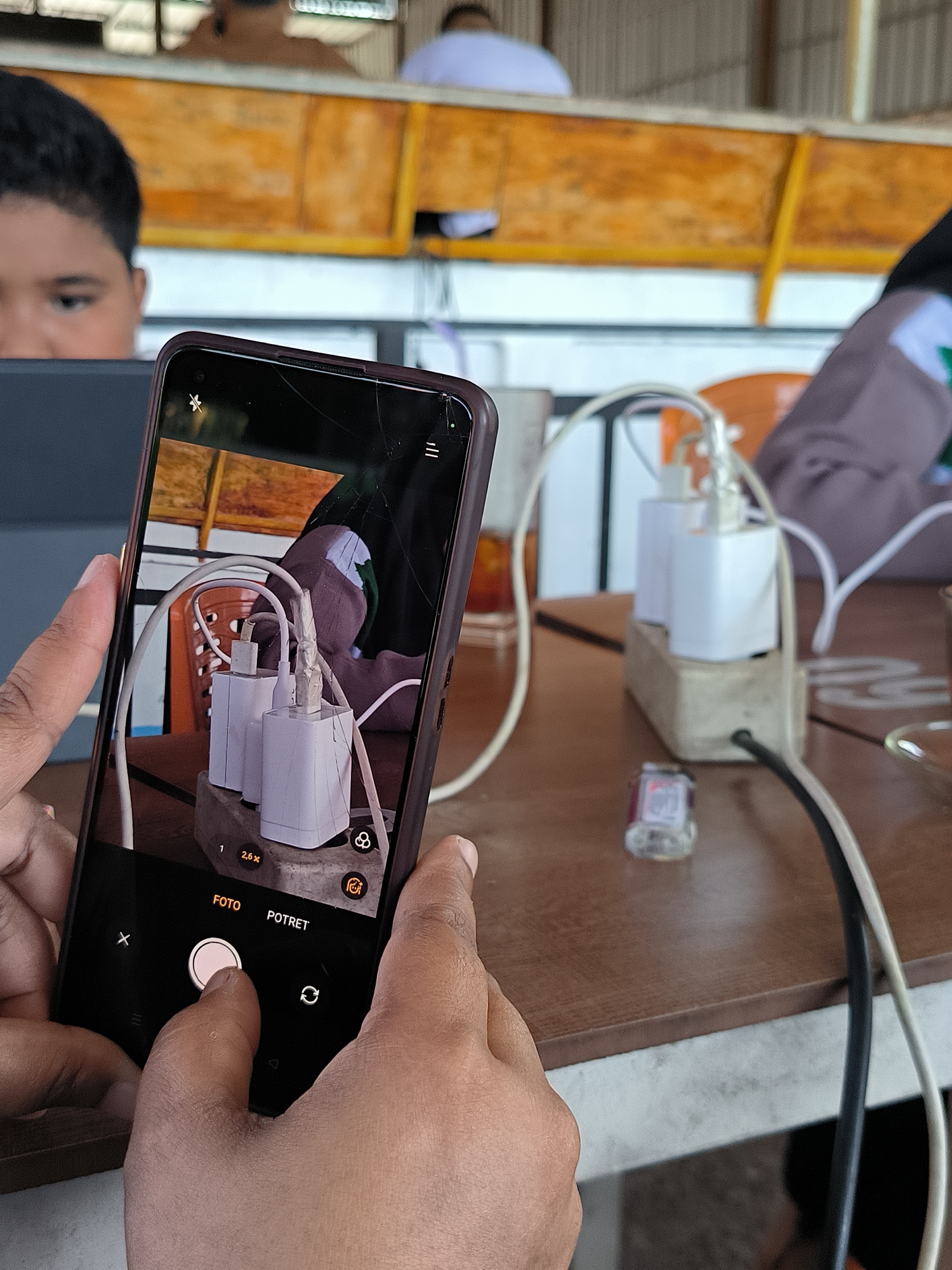 A person is taking a photo with a smartphone of an electrical outlet with two white USB chargers plugged into it on a wooden table. A small, clear bottle with a red label is next to the outlet. Cords from the chargers are visible. In the background, there is a person behind a laptop screen, another person seated near the outlet, and two people sitting or standing at a counter or table further back. There are cracks visible on the screen of the smartphone. The camera is in "Foto" mode. The location is presumably Matang, Indonesia.