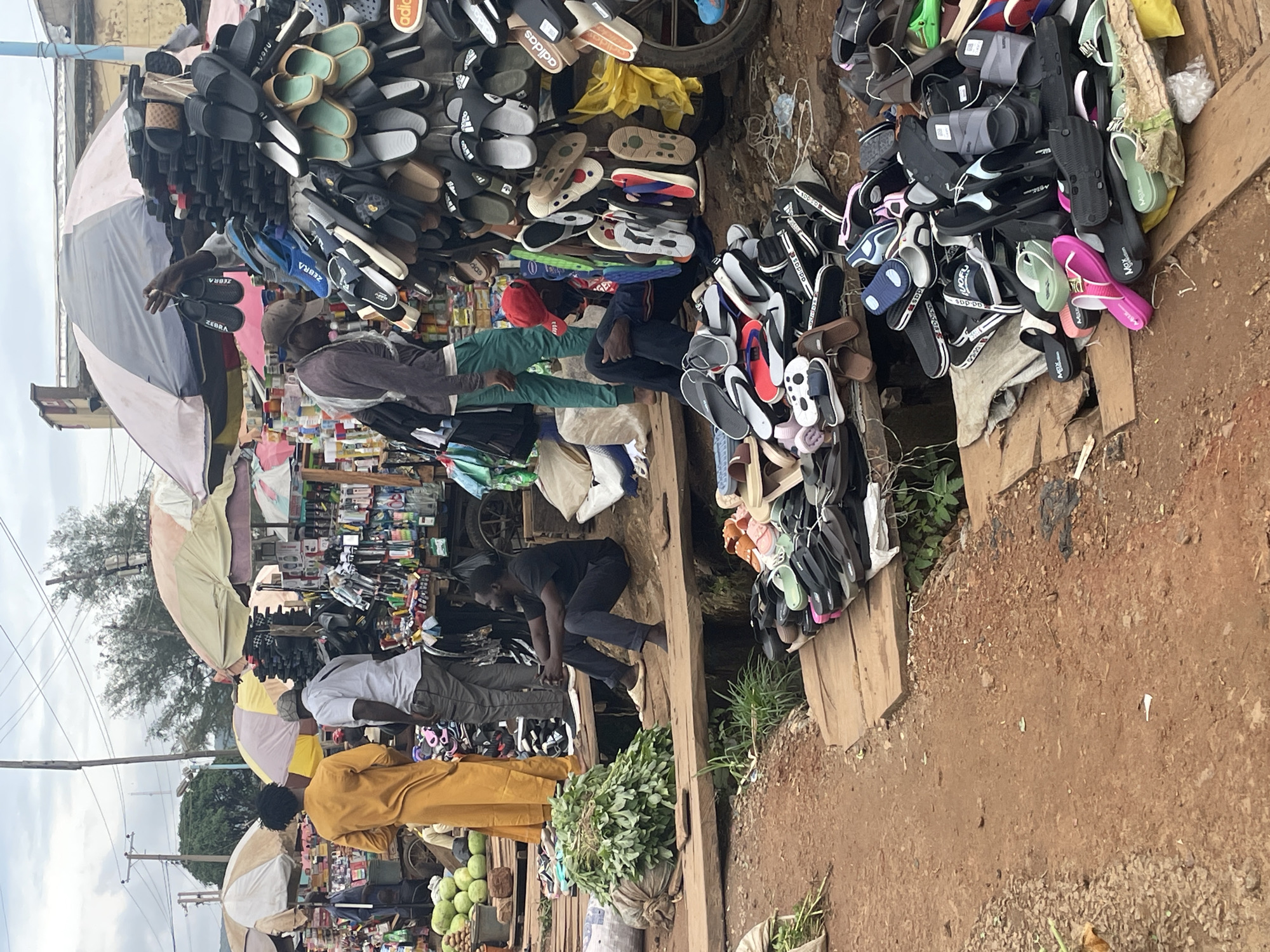 This media file shows a street market scene in Rafin Par, Nigeria. A variety of footwear, primarily flip-flops and sandals, are displayed on wooden shelves and tables. The shoes are in numerous colors and styles, with some bearing brand names like "Adidas" and "Max". People are seen browsing and working at the market. One individual is tending to the footwear display, while others are interacting with vendors or walking through the market.
In the foreground, a person wearing a yellow outfit is visible. Further back, another person in a grey shirt and dark pants is seen. The market appears to be outdoors, with a dirt ground and some greenery visible. The sky suggests it may be daytime, possibly overcast. There are also some umbrellas, suggesting a need for shade or protection from potential rain.
The scene conveys a bustling market atmosphere, with goods piled high and people engaged in commerce. The presence of food items, such as fruits, indicates a diverse market offering. The overall impression is that of a lively local marketplace in Nigeria.