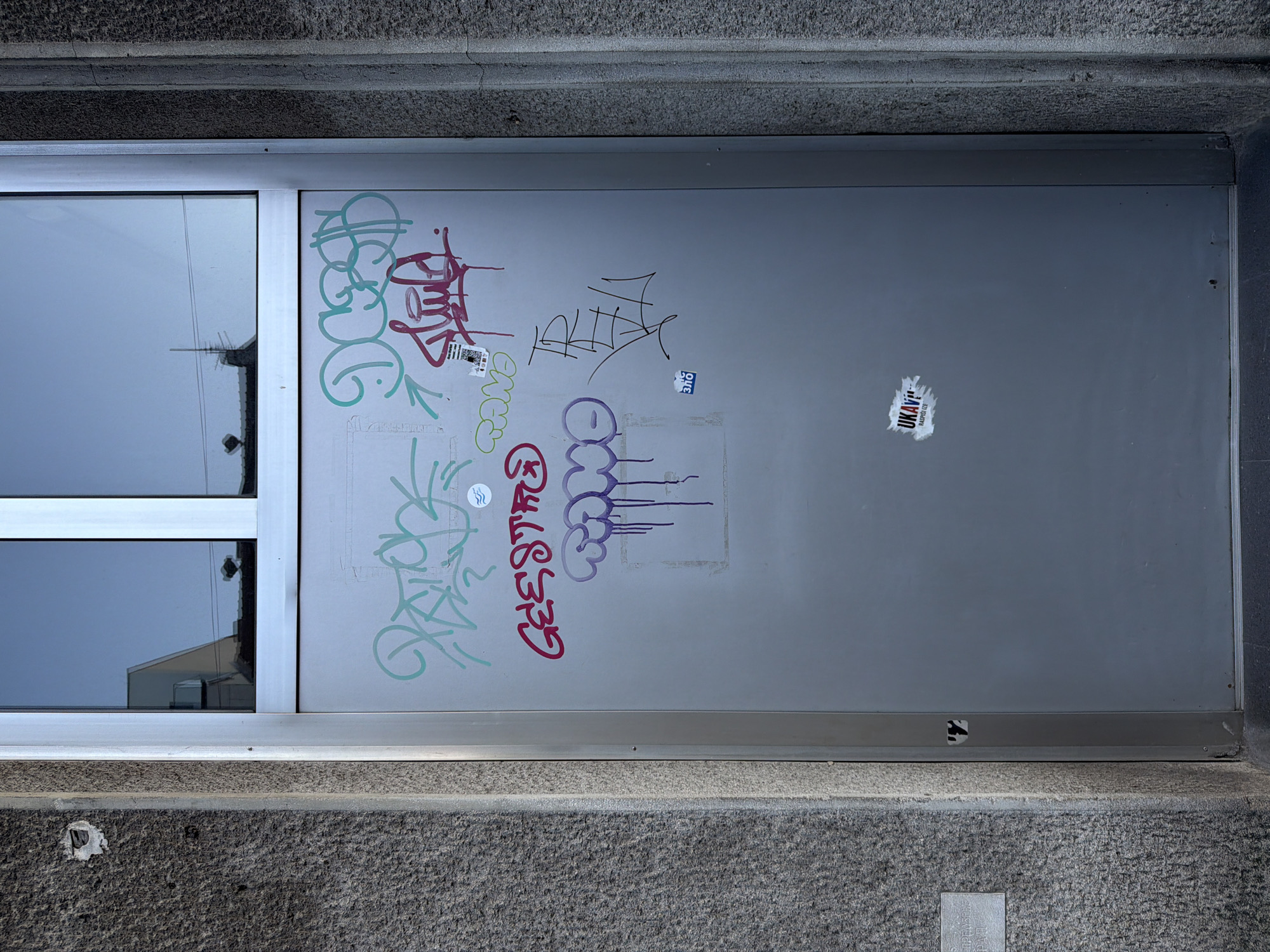 A ground-level view shows a closed window and gray metal door heavily marked with graffiti. The window reflects the sky and a nearby building. The graffiti consists of illegible tags in various colors, including green, red, purple, and black. Stickers are also affixed to the door. The door is set within a gray, rough-textured stone or concrete frame. The location is Belgrade, Serbia.
