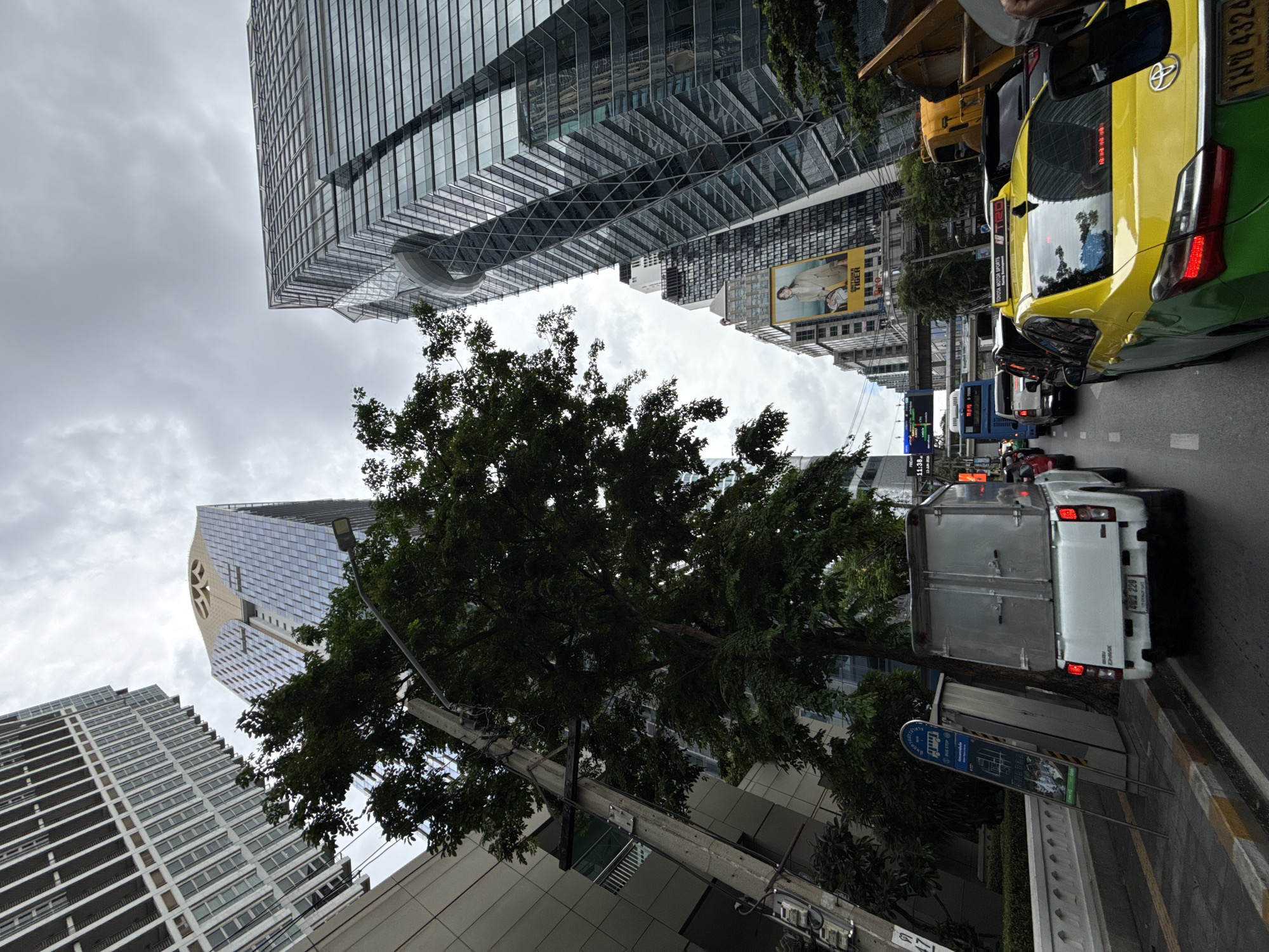 This is a street-level, eye-level, vertical shot of a traffic jam on a cloudy day in Bangkok, Thailand. The perspective is looking forward along a city street lined with tall, modern buildings. The prominent foreground elements are the rear of a white utility truck and the rear of a yellow and green taxi. The taxi has a license plate that reads "1กผ 4324". The utility truck has a license plate that reads "บข 228". In the middle ground, there are more vehicles, including a blue bus, and further down the street, a large billboard features a woman in a tan jacket. The background is dominated by the towering glass and steel facades of skyscrapers, reflecting the overcast sky. A large green tree stands to the left of the frame, partially obscuring the buildings. There is a bus stop sign to the left of the utility truck. The overall scene depicts urban congestion in a bustling metropolitan area.