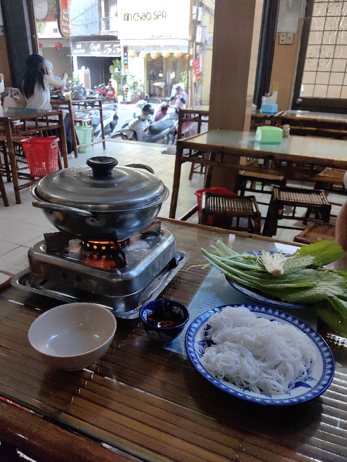 The image captures an indoor dining area of a restaurant in Thuận Hóa Ward, Vietnam, with a direct view of an urban street beyond. In the foreground, a metallic hot pot with a lid is centered on an activated portable stove, exhibiting an orange flame. To the right, on a bamboo-slat table, a blue and white patterned plate holds a portion of white rice noodles. Adjacent to the noodles, another blue and white patterned plate contains an assortment of fresh green leafy vegetables and enoki mushrooms. A small, dark blue bowl with a red substance, likely chili flakes or sauce, and an empty light-colored ceramic bowl are also present on the table.
In the midground, several wooden tables and stools are arranged within the restaurant's interior. A person with long dark hair, wearing a white top, is seated at a table to the far left, partially visible. A red plastic basket is positioned beneath a table. The background displays a busy street scene with multiple motorbikes, both parked and in motion. Commercial storefronts line the street, including one labeled "XIN CHAO SPA" and another identifiable as "Cafe 55." Several indistinct figures are visible on the street.