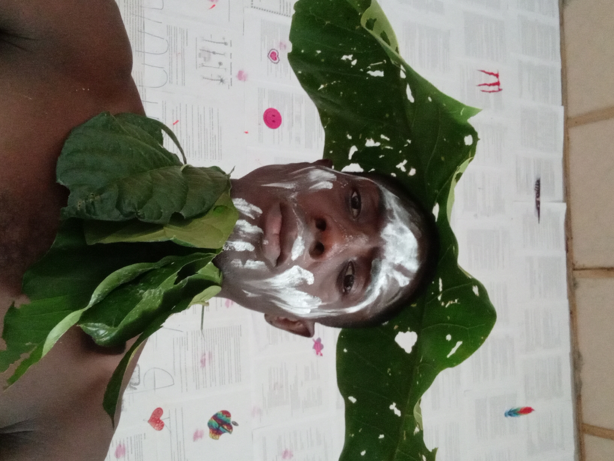 A close-up, slightly angled shot shows a man's upper body. His face is painted with white markings. Green leaves are arranged around his neck and head. The background is a white surface with drawings, lettering, and small decorative images. The location is presumed to be Akwete, Nigeria.