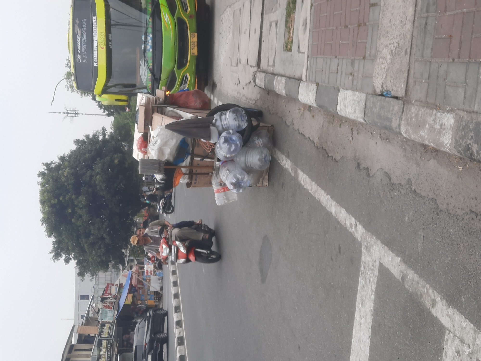 In North Jakarta, Indonesia, a street scene unfolds with a green and yellow bus, branded "JETBUS PARIWISATA," parked on the right side. In front of the bus, a cart loaded with water bottles and various other items is positioned. A man wearing a yellow cap and a patterned shirt rides a red motorcycle with a passenger behind him, traveling down the street from left to right. To the left of the street, a black car is parked in front of a row of buildings. The environment is characterized by an overcast sky, a paved road with white markings, and a sidewalk.