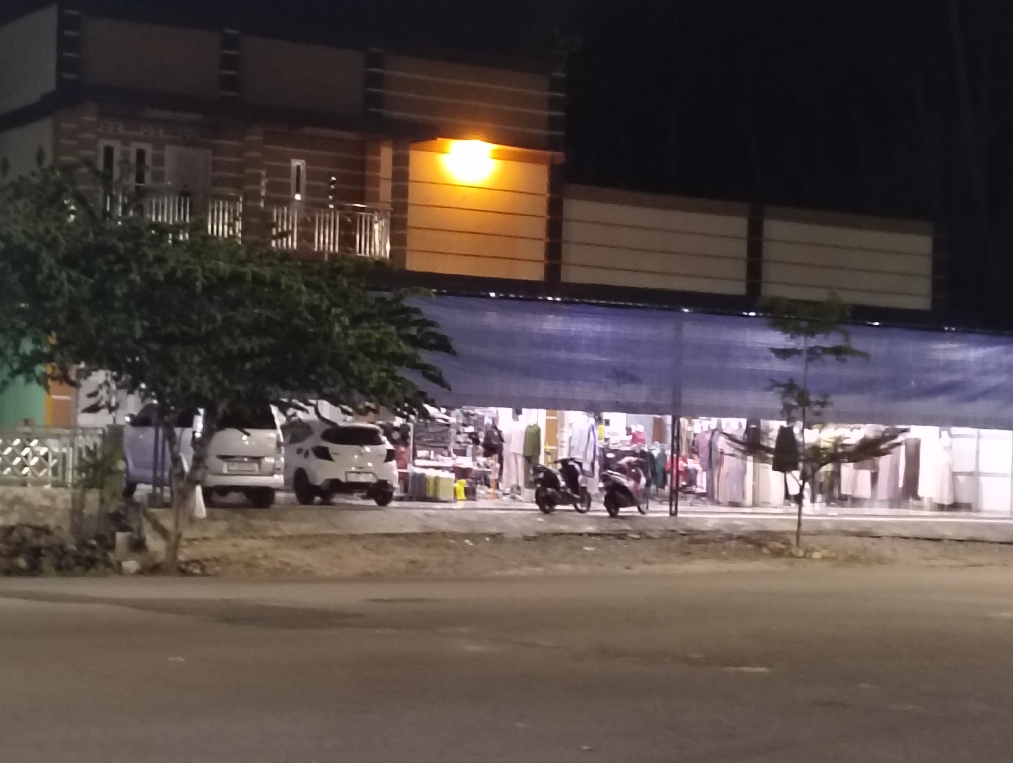 In Kampal, Indonesia, at night, a street-level view captures a building with a brown brick facade and horizontal cream-colored siding, illuminated by an orange outdoor light. A balcony with white railings is visible on the upper level. Below, a row of shops is set up behind a blue tarp. Two white cars are parked in front of the shops, and two motorcycles are positioned near the entrance. The ground in front of the shops appears to be a dirt or gravel lot, with a paved road in the foreground. The scene is dark, with the artificial lights providing the primary illumination.