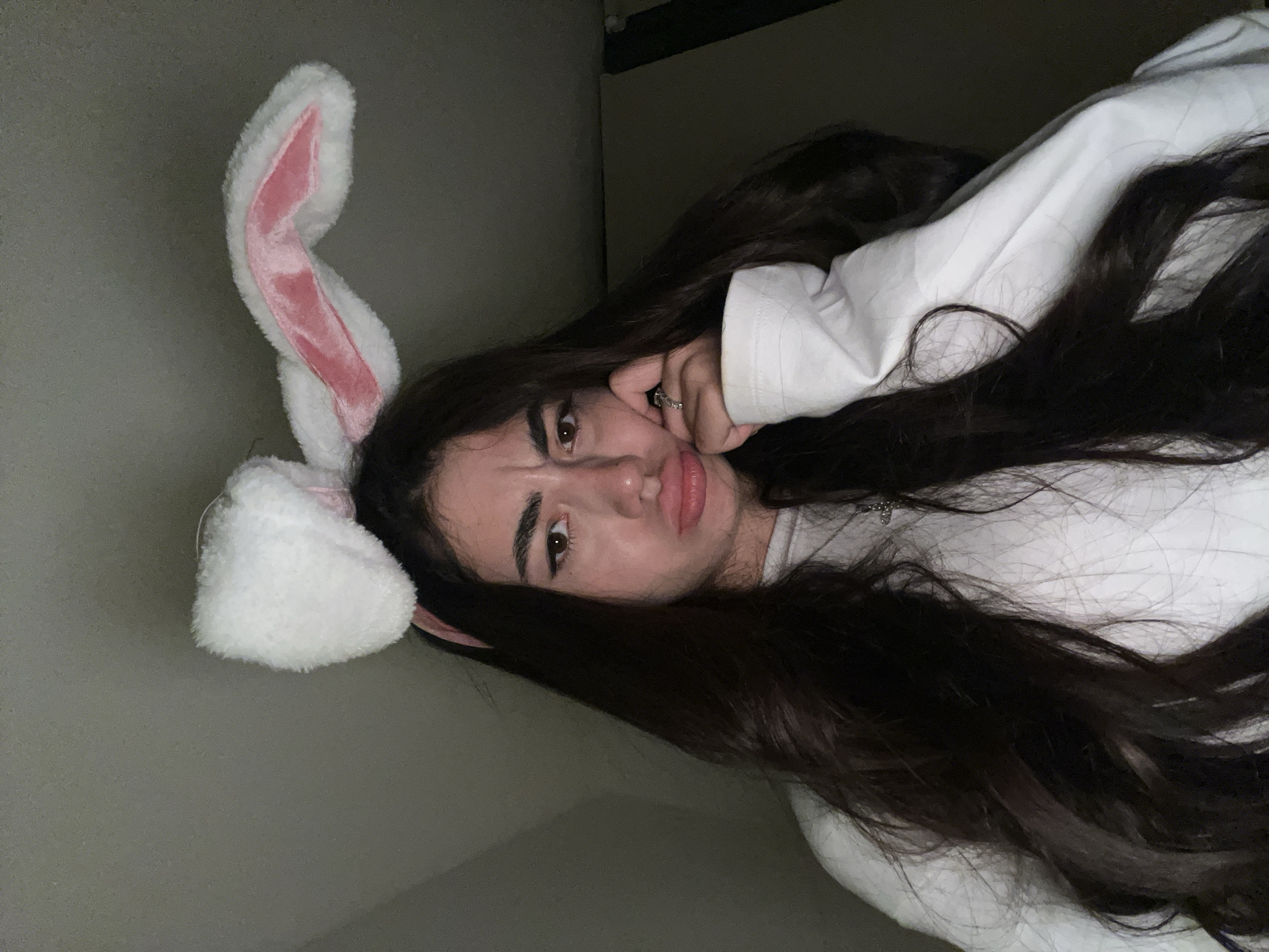 A young woman with long, dark hair wears a white fluffy headband with pink bunny ears. She is leaning her head on her hand, with her fingers touching her chin and cheek, and her lips are pursed. She is wearing a white long-sleeved shirt and a necklace. The background is dark and appears to be an indoor setting with a wall. There are no discernible features to identify the location as Chotěšov, Czechia.